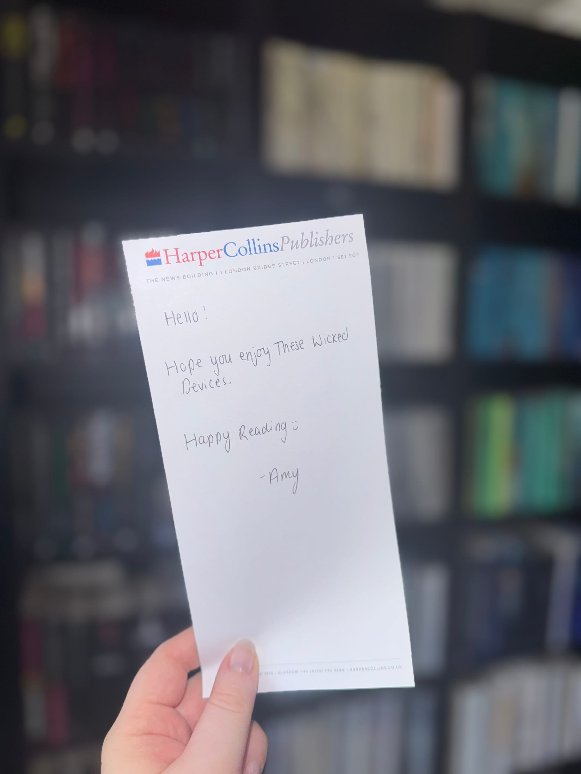 Hand holding a HarperCollins note reading, "Hello! Hope you enjoy These Wicked Devices. Happy Reading :) -Amy," with blurred bookshelves in the background.