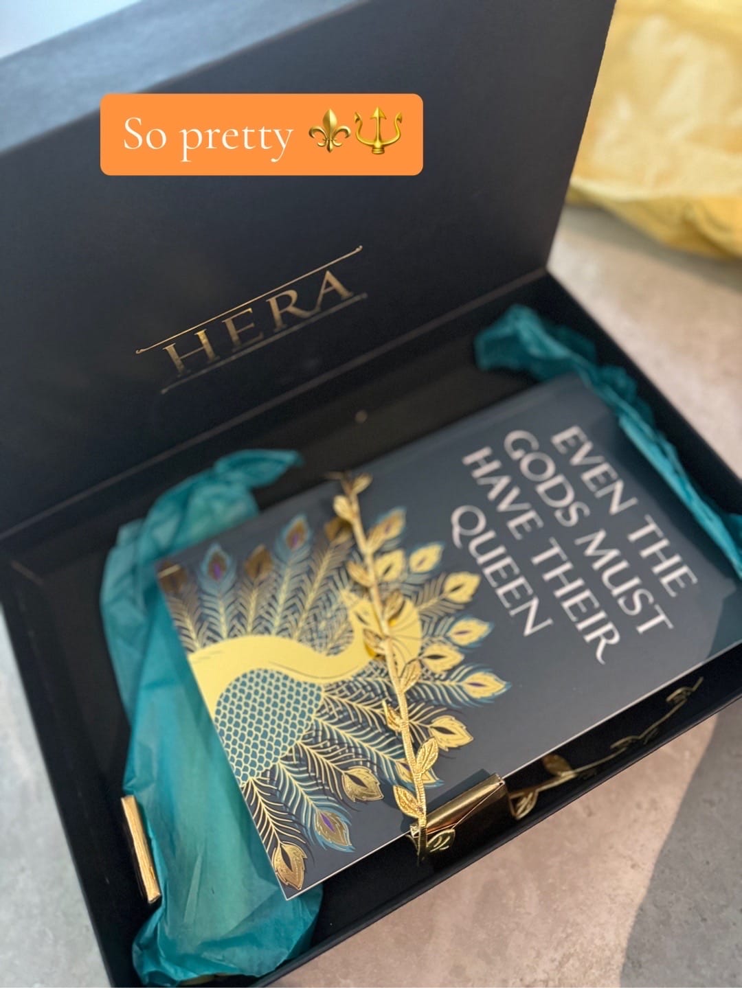 A black HERA box contains a decorative book with "EVEN THE GODS MUST HAVE THEIR QUEEN" text, gold peacock design, and teal wrap.
