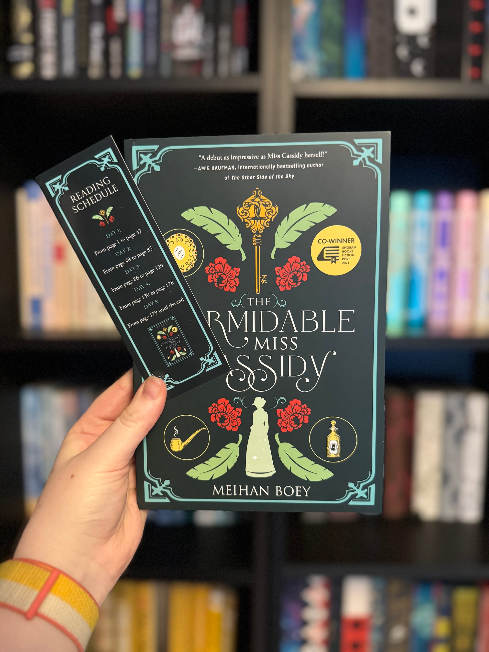 Hand holds "The Formidable Miss Cassidy" book and reading schedule bookmark. Dark cover with floral design. Bookshelf blurred in background.
