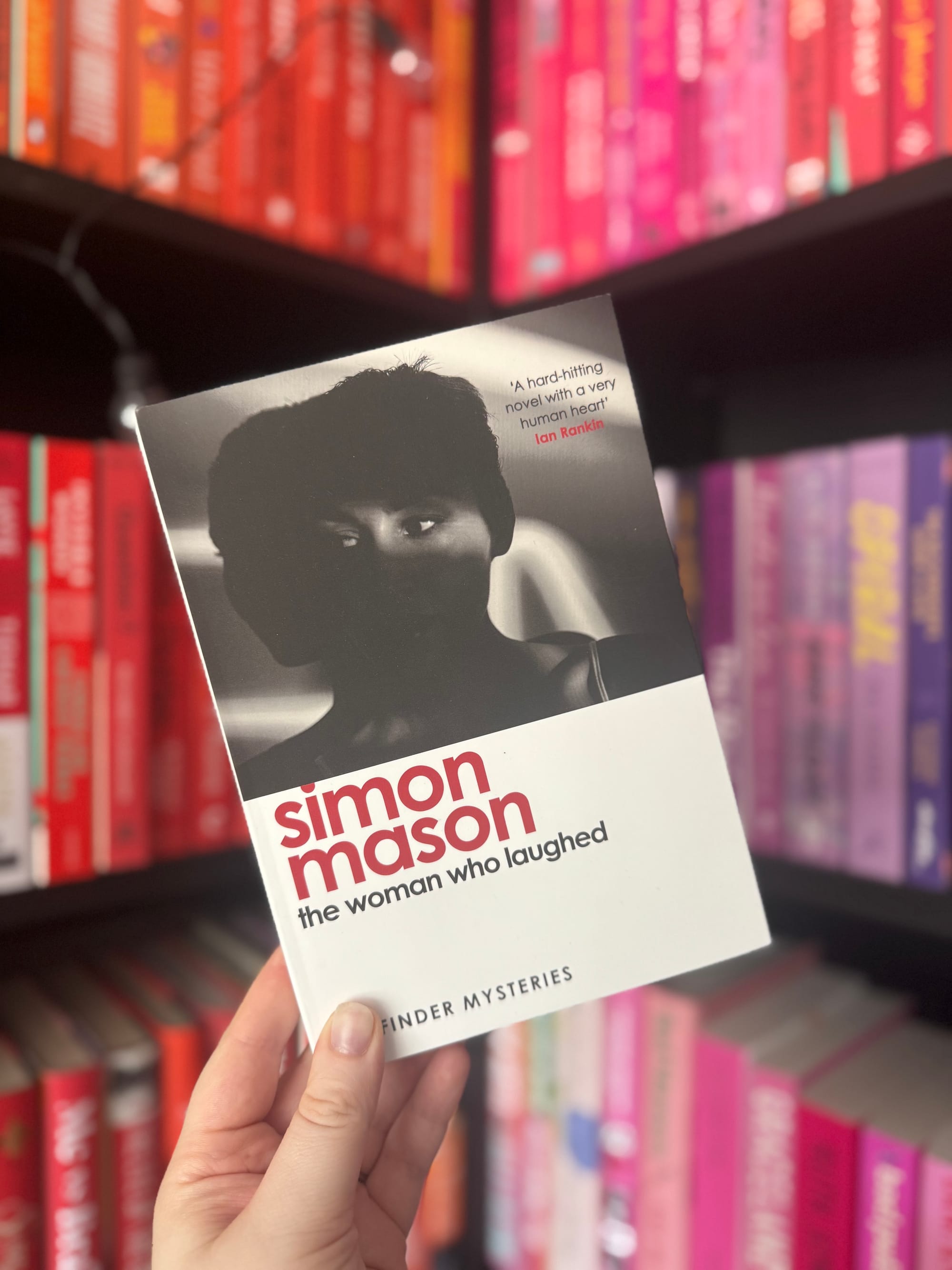 Hand holding a book titled "The Woman Who Laughed" by Simon Mason. Colorful bookshelves in the background create a vibrant setting.