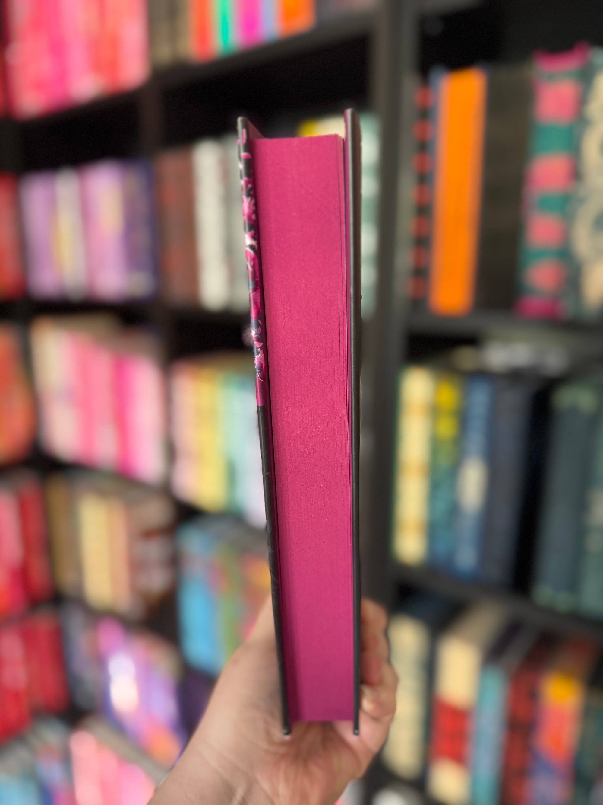 Hand holding a book with a pink edge, in front of a colorful bookcase. Books display a variety of bright, vivid covers.