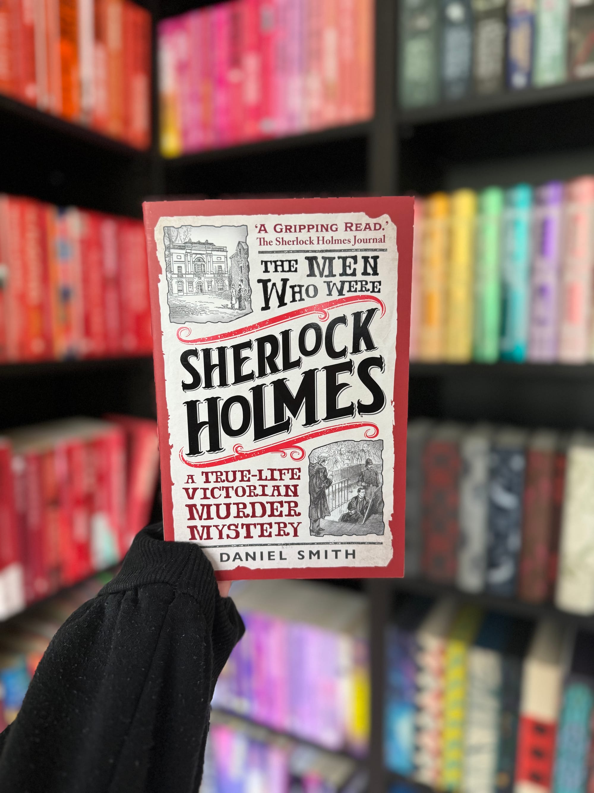 Hand holds "The Men Who Were Sherlock Holmes" book by Daniel Smith. Background: colorful bookshelf. Book has red and black text.