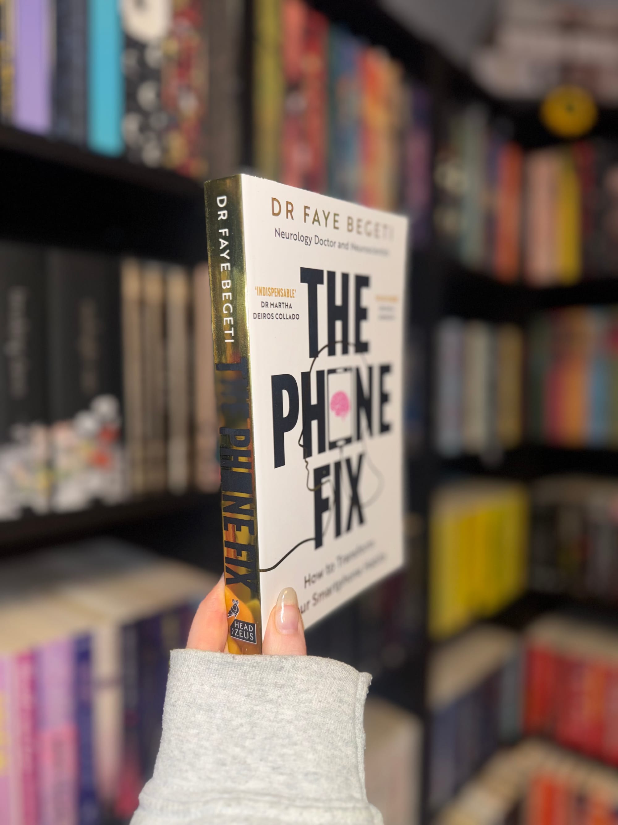 Person holds "The Phone Fix" book by Dr Faye Begeti in a library. The background is blurred, featuring colorful books on shelves.