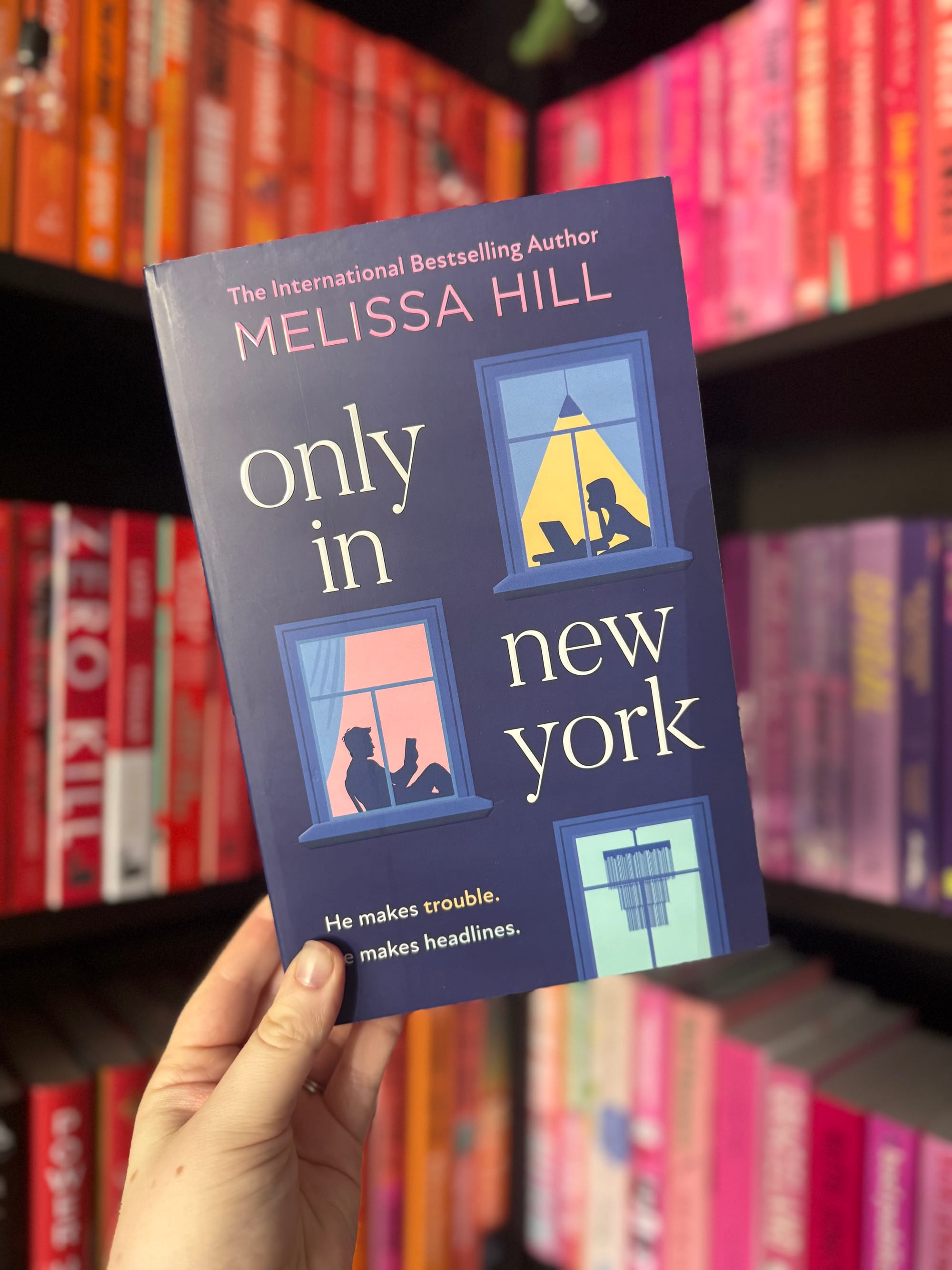 A hand holds "Only in New York" by Melissa Hill. The cover shows silhouettes of people at windows. Background: colorful bookshelves.