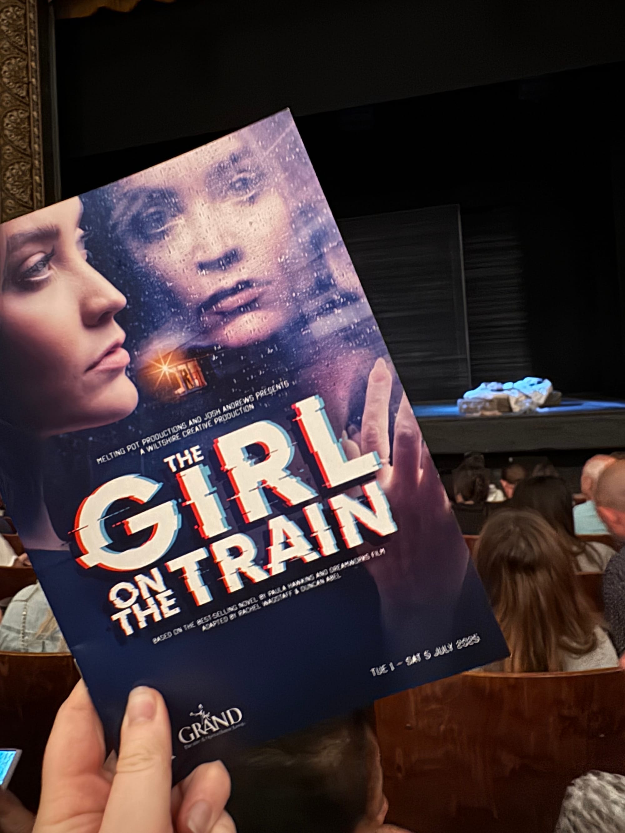 Hand holding "The Girl on the Train" program in a theater, with stage set in background. Moody cover art visible, audience seated.