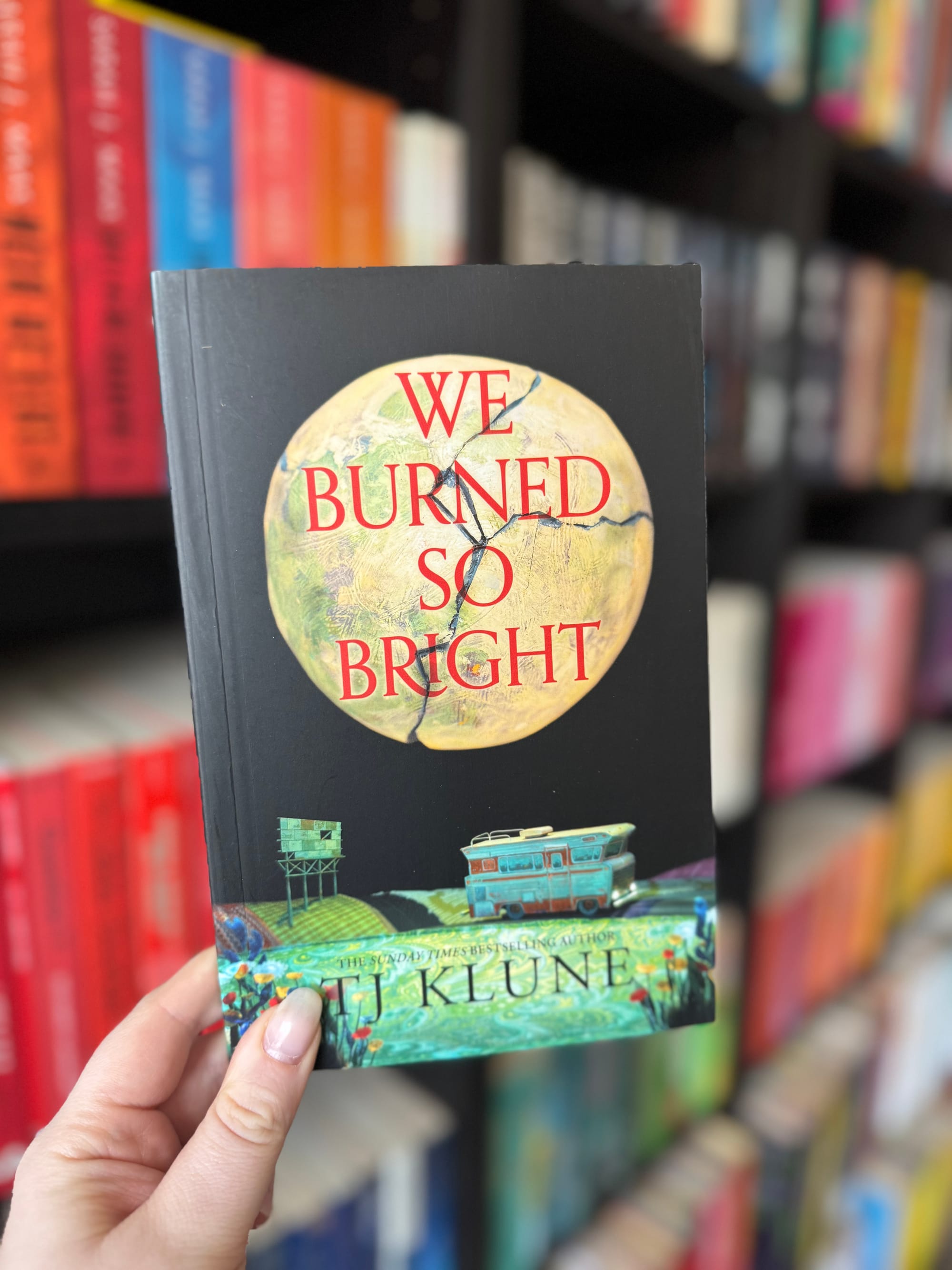 Hand holding a book titled "We Burned So Bright" by TJ Klune. Background has colorful books on shelves, creating a vibrant bookstore setting.