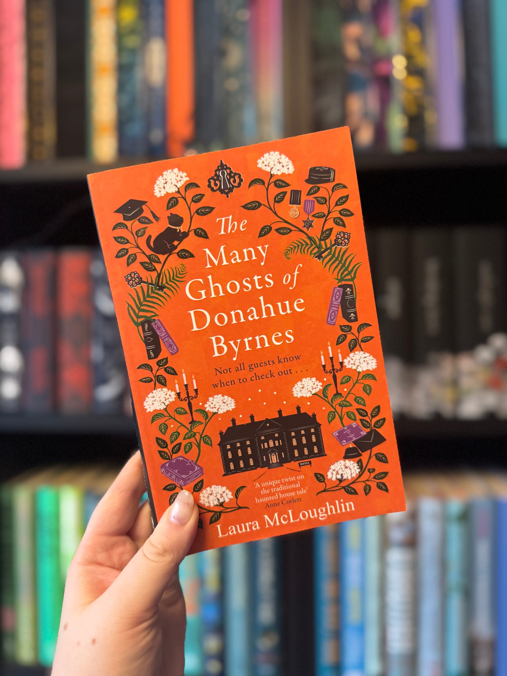 A hand holds "The Many Ghosts of Donahue Byrnes" book with an orange cover, floral design, and text. Bookshelf background is blurred.