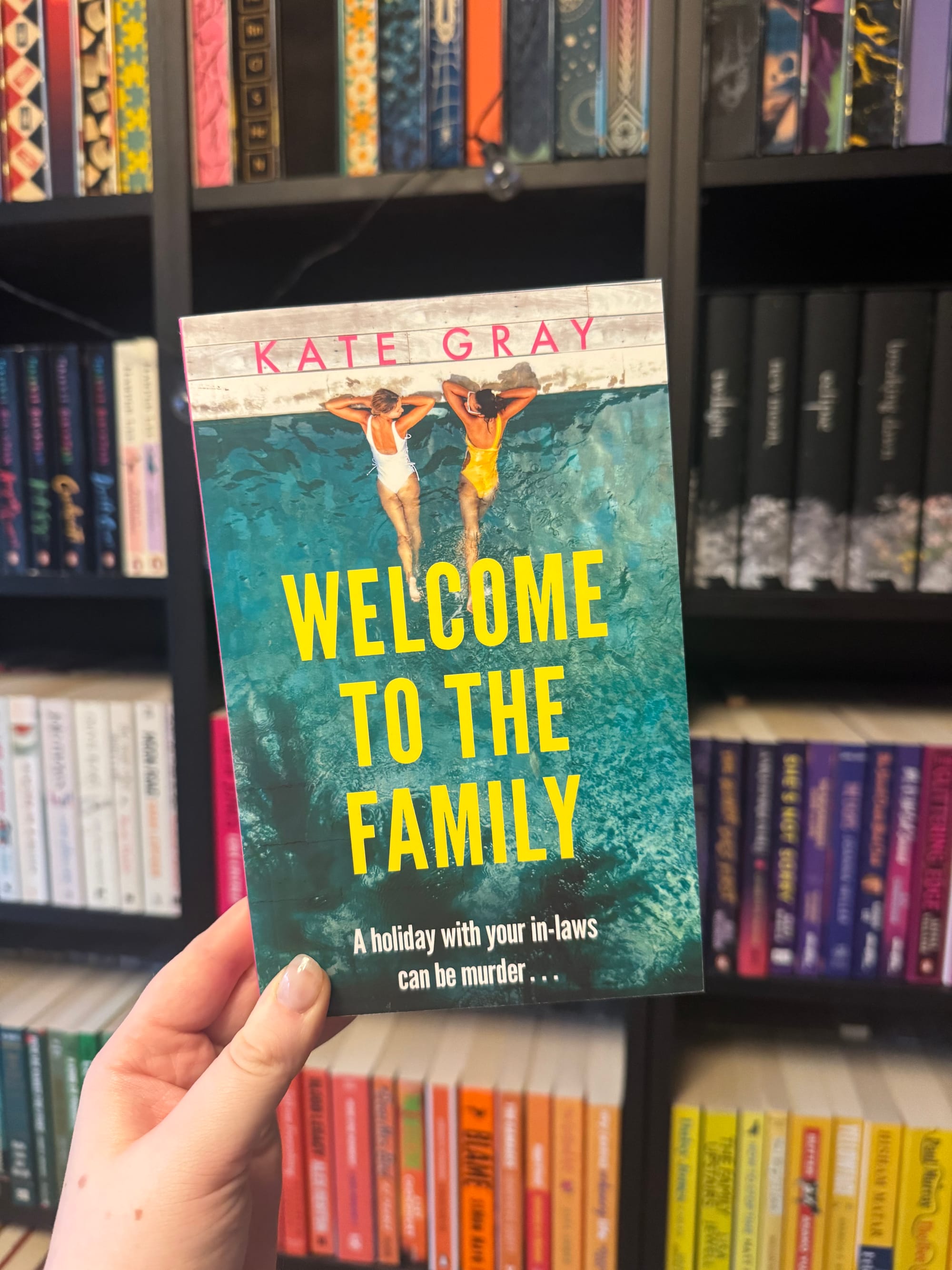 Hand holding the book "Welcome to the Family" by Kate Gray, set against colorful bookshelves. Two people in swimsuits on the cover, poolside.