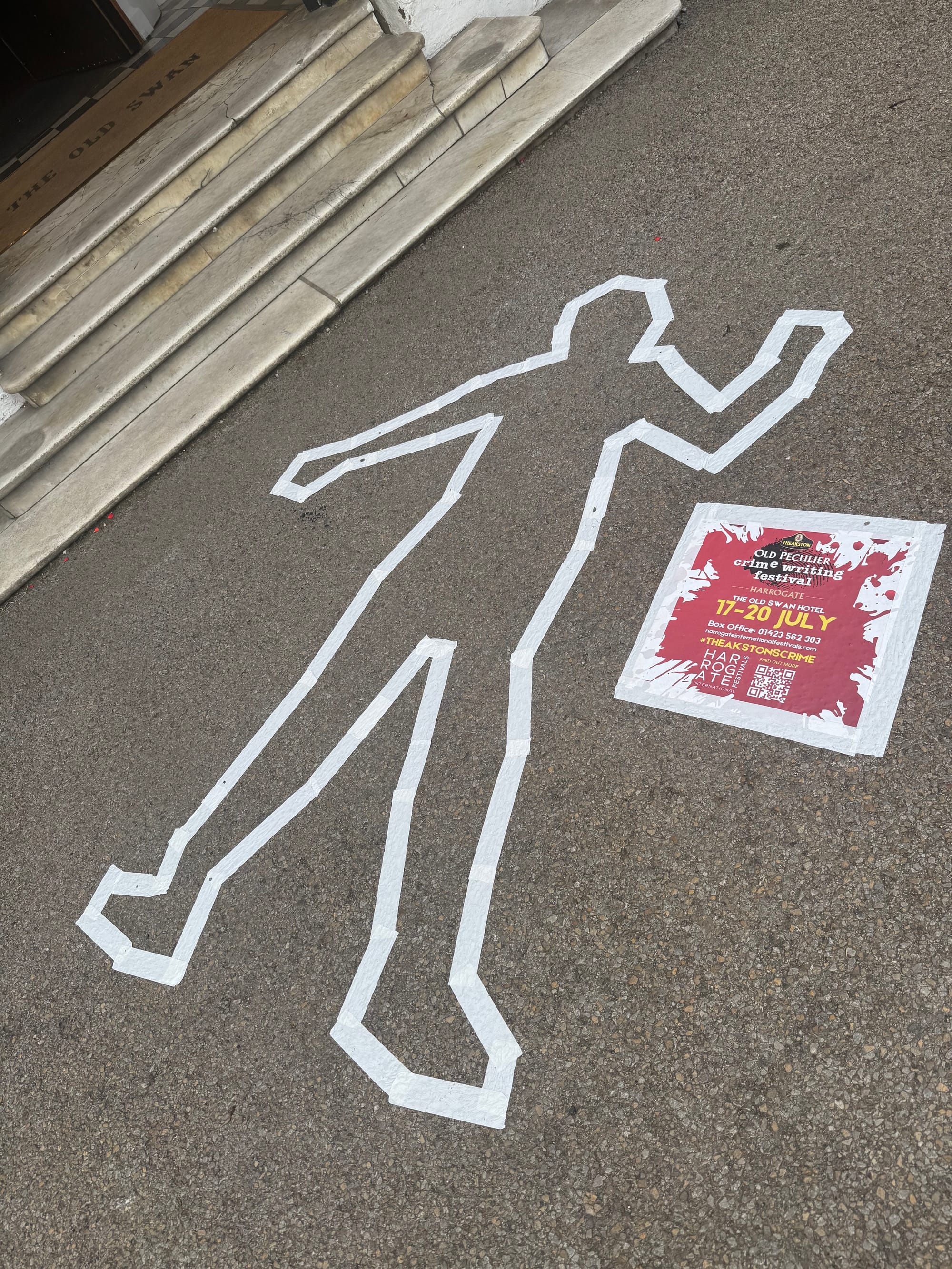 A white tape outline of a human figure on the pavement near steps, next to a colorful event poster promoting a murder mystery show in July.