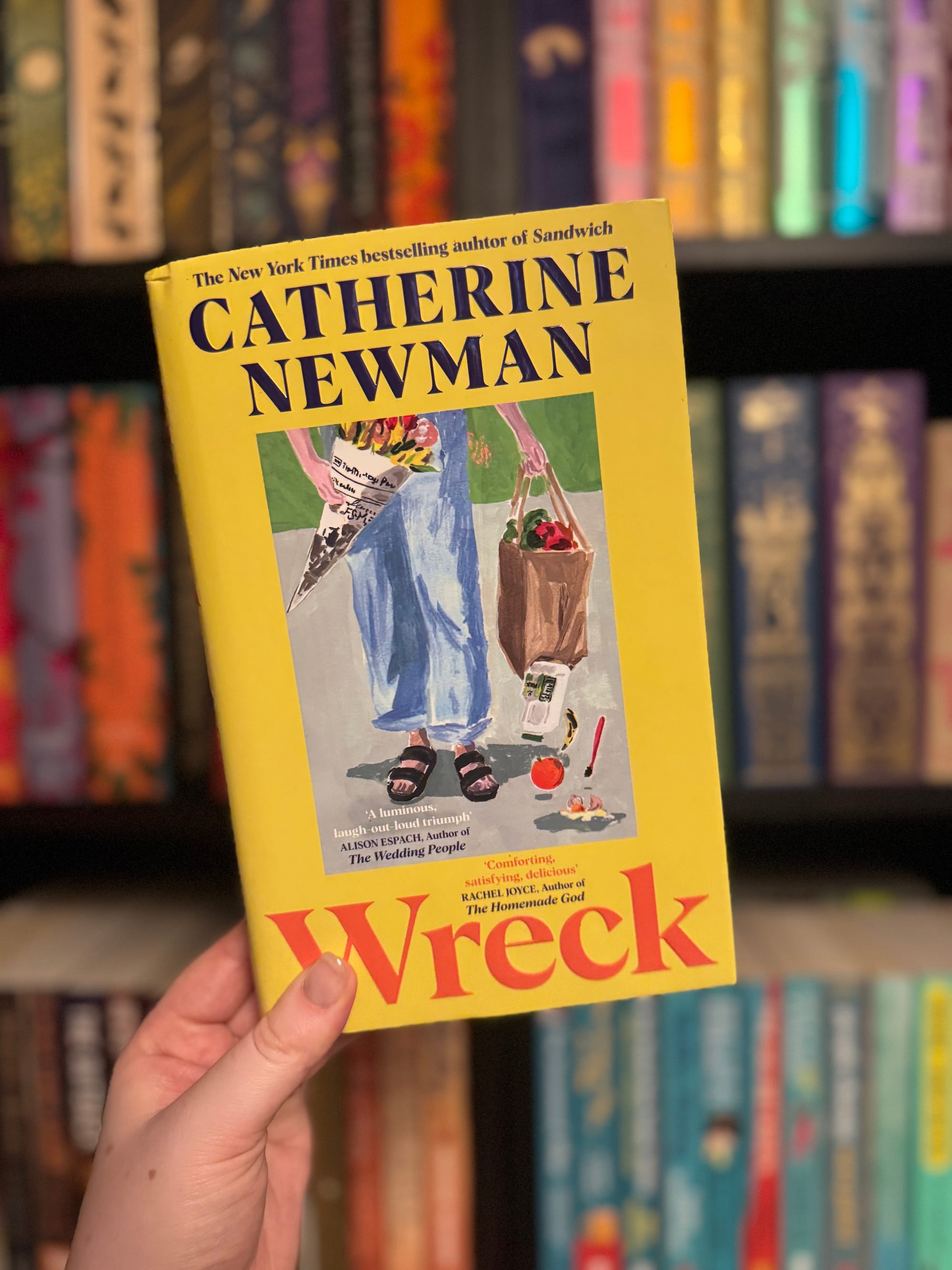 A hand holds a yellow book titled "Wreck" by Catherine Newman. The book shows a painting with groceries. Blurred colorful books in the background.