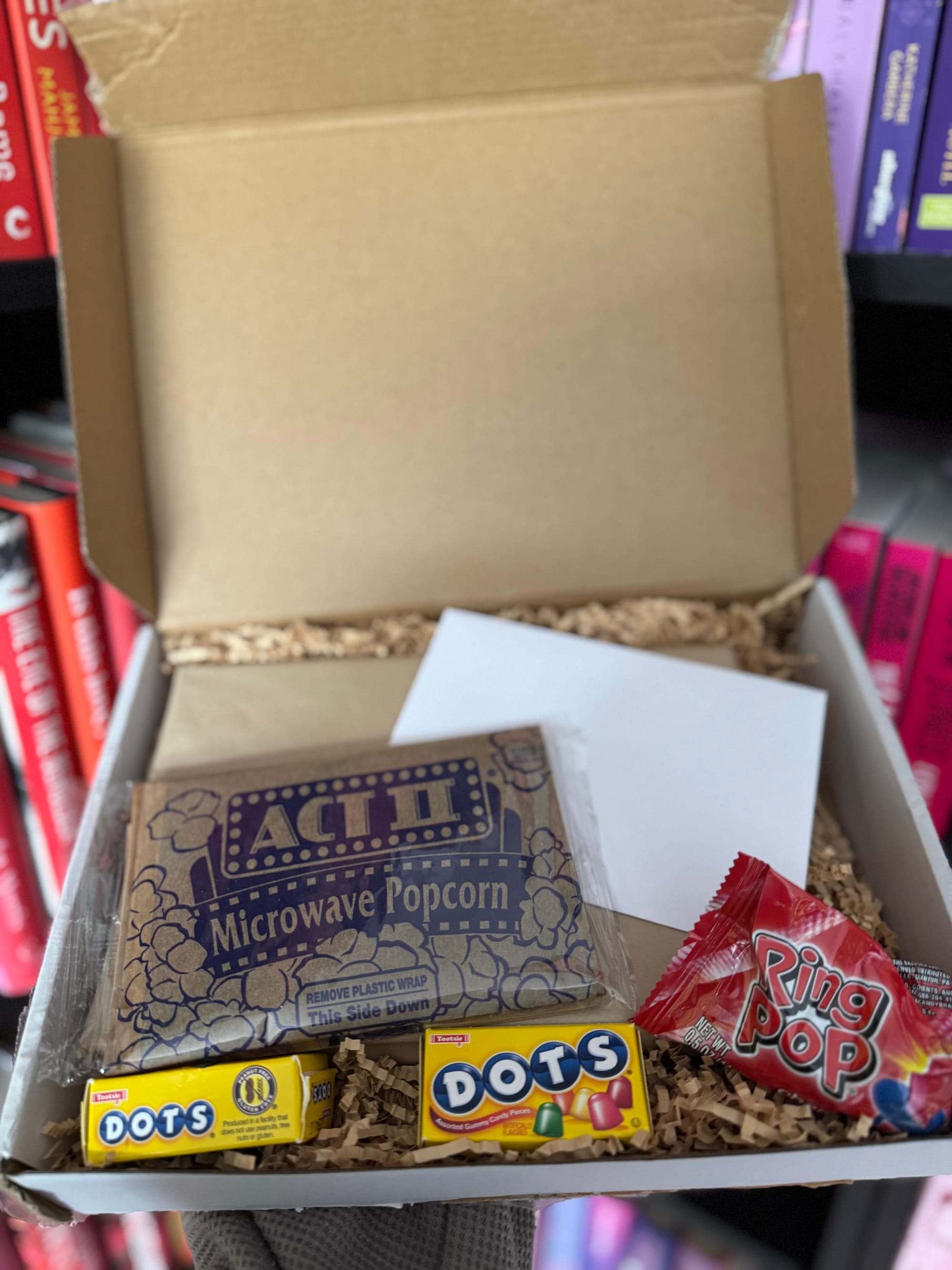 An open gift box filled with shredded paper contains a packet of ACT II Microwave Popcorn, two boxes of Dots candy, a Ring Pop, and a blank white envelope. The box is being held in front of a colourful bookshelf arranged by spine colour, featuring red, pink, and purple books.