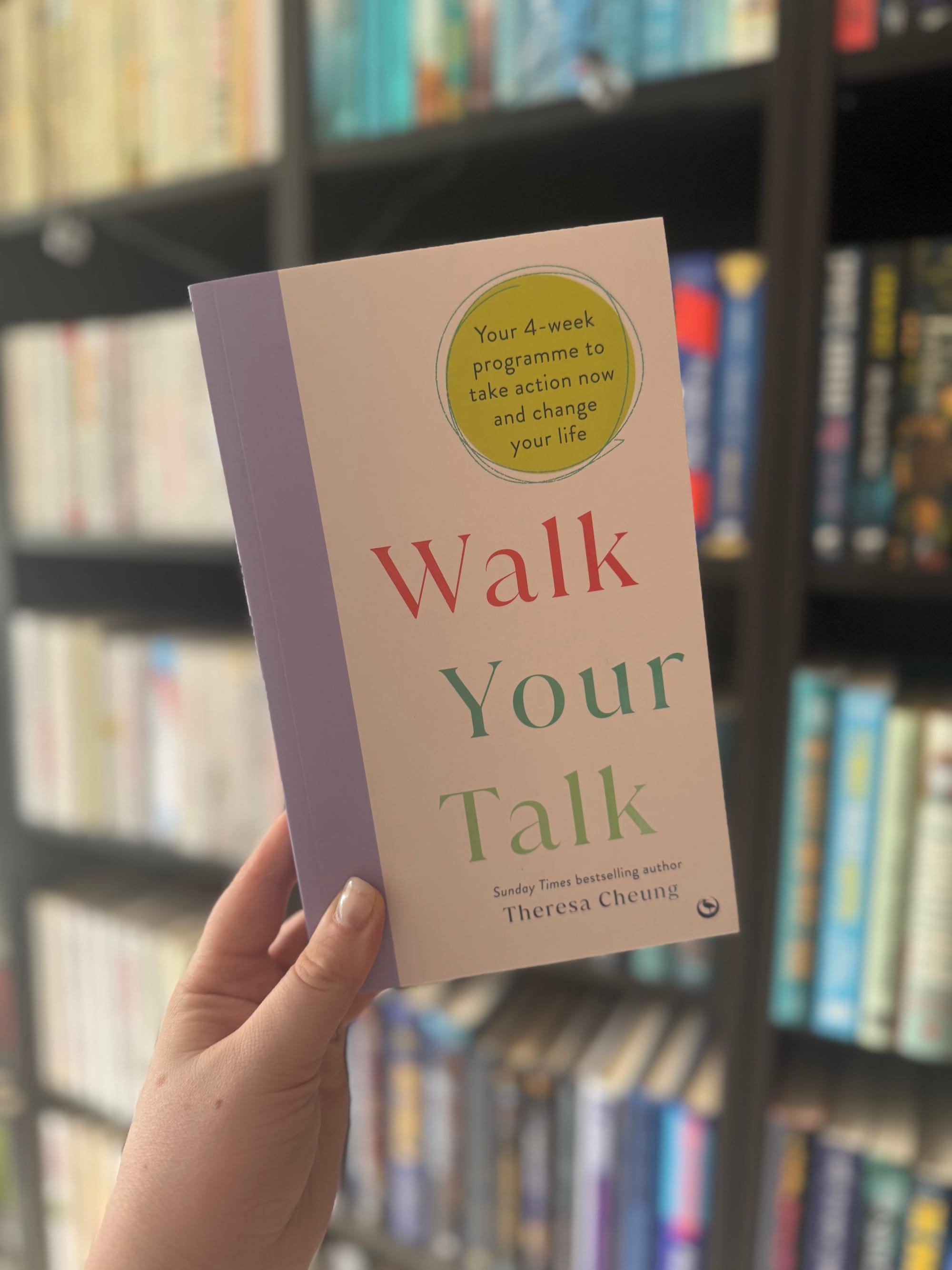 Hand holding a book titled "Walk Your Talk" in front of a blurred bookshelf. The cover has a green and purple design.