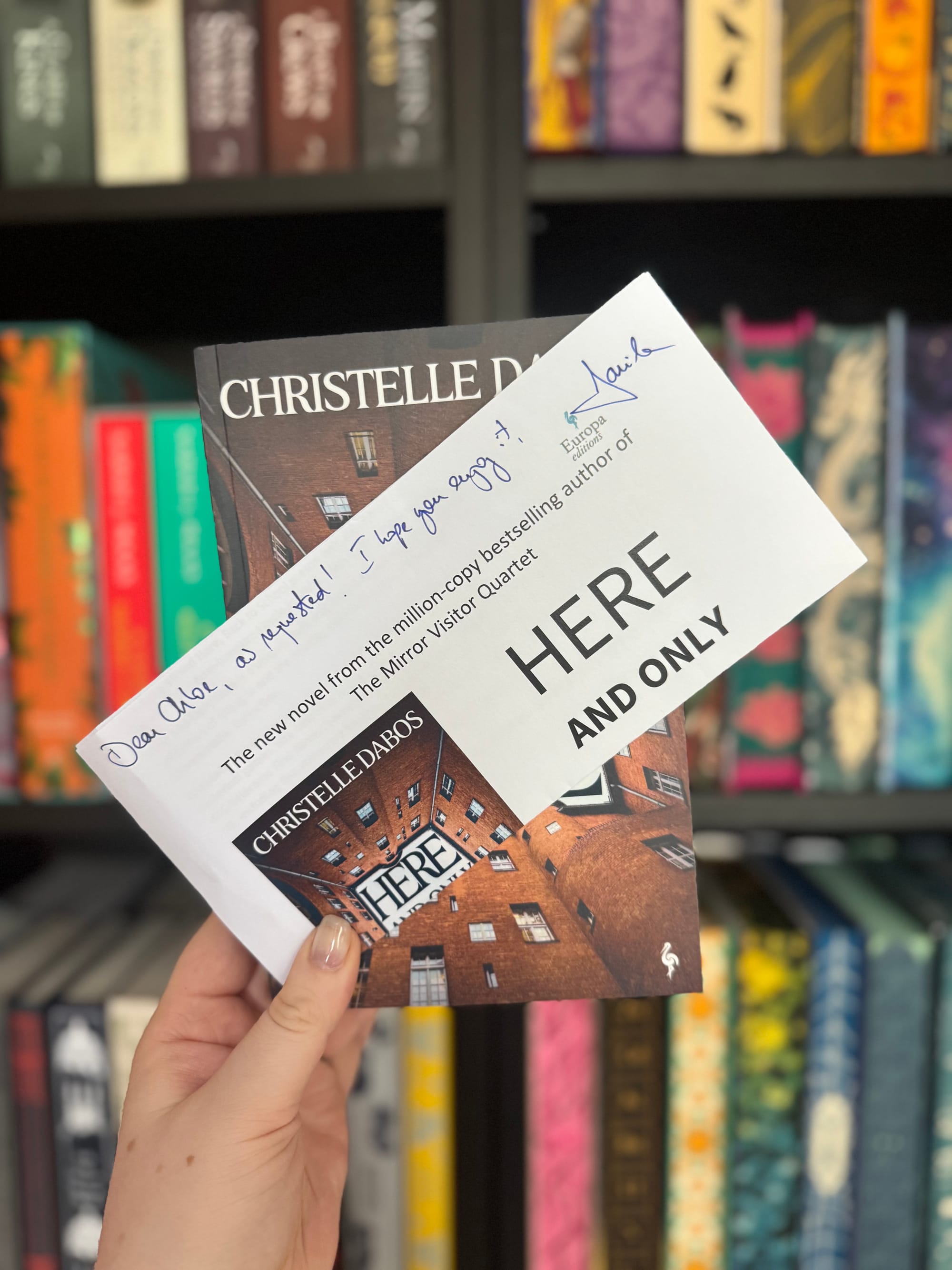 A person holds the book Here and Only Here by Christelle Dabos in front of a colourful bookshelf. A white note with blue handwritten text is partially covering the book’s cover, which shows a brick courtyard with the title written across the centre.