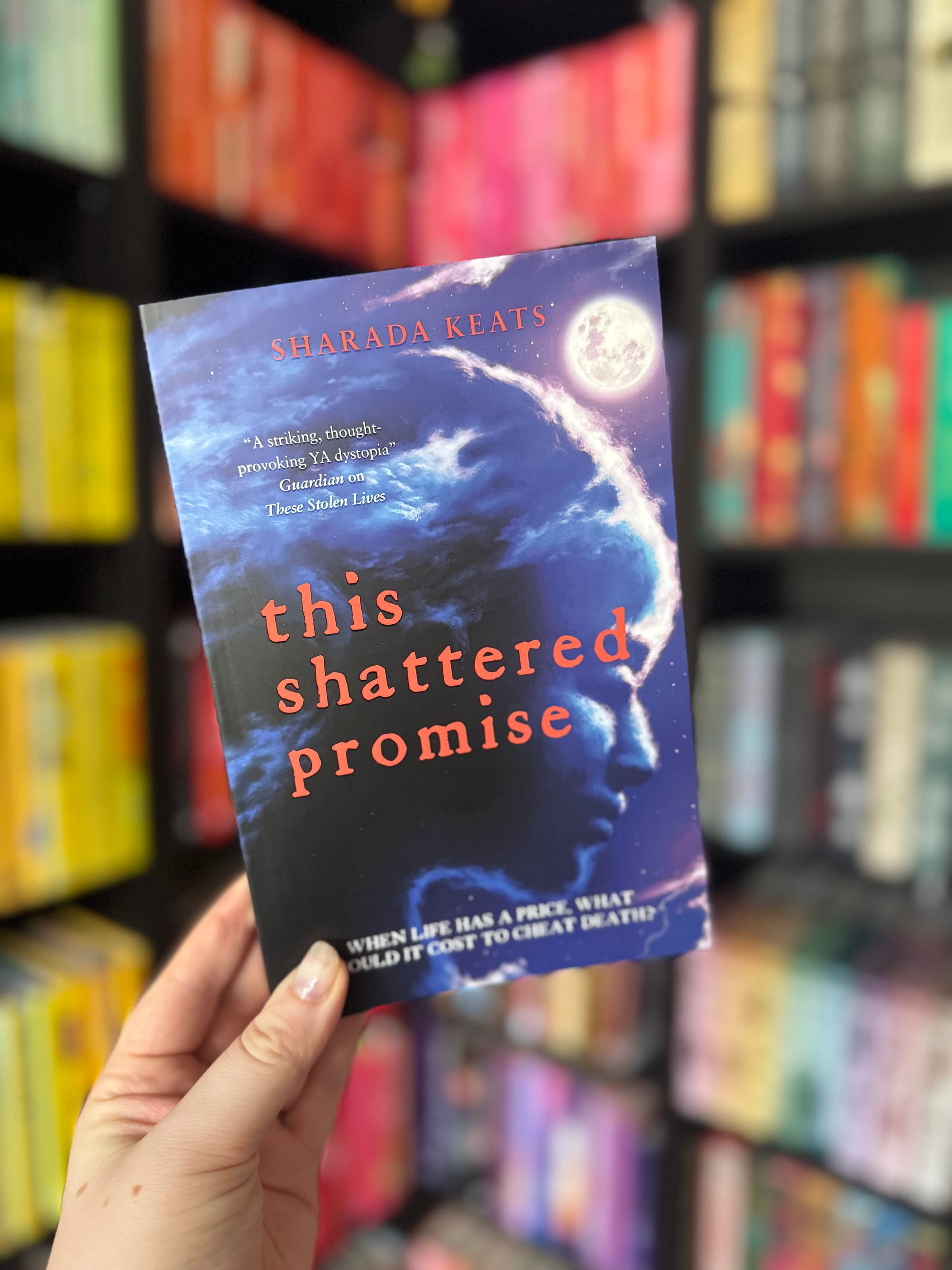Hand holding a book titled "This Shattered Promise" by Sharada Keats against a colorful bookshelf. The cover shows a face with cosmic imagery.