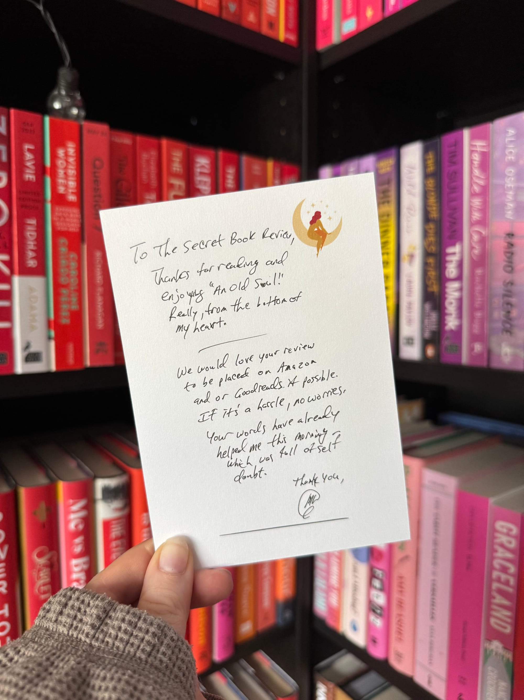 A hand holds a handwritten note in front of a colour-coordinated bookshelf with red, pink, and purple spines. The note is addressed to "The Secret Book Review" and thanks them for reading and enjoying An Old Soul. It includes a request to post the review on Amazon or Goodreads, if possible. The message is signed with a small drawing and initials.