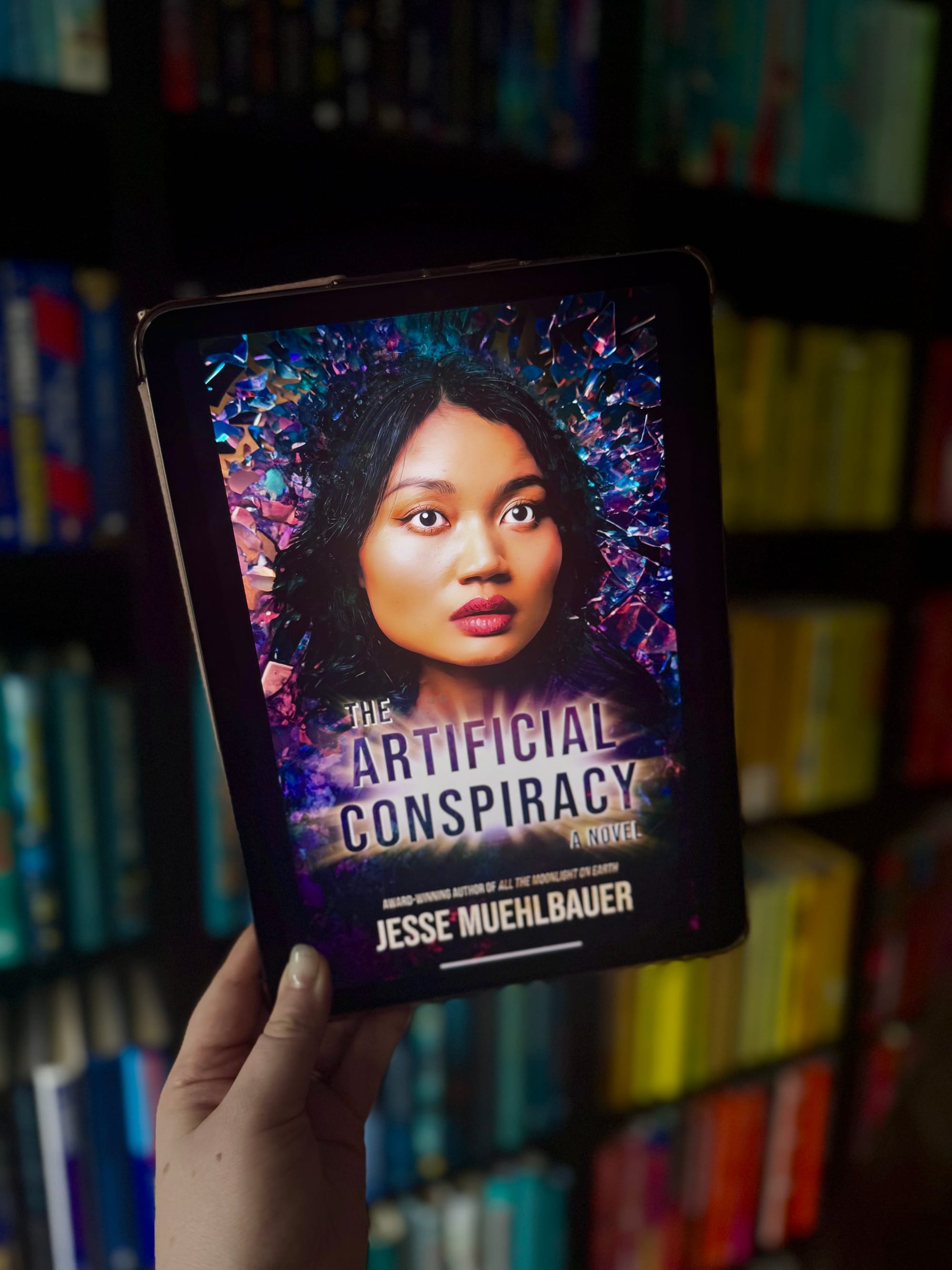 A hand holds a tablet displaying a book titled "The Artificial Conspiracy" by Jesse Muehlbauer, against a colorful, blurred bookshelf background.