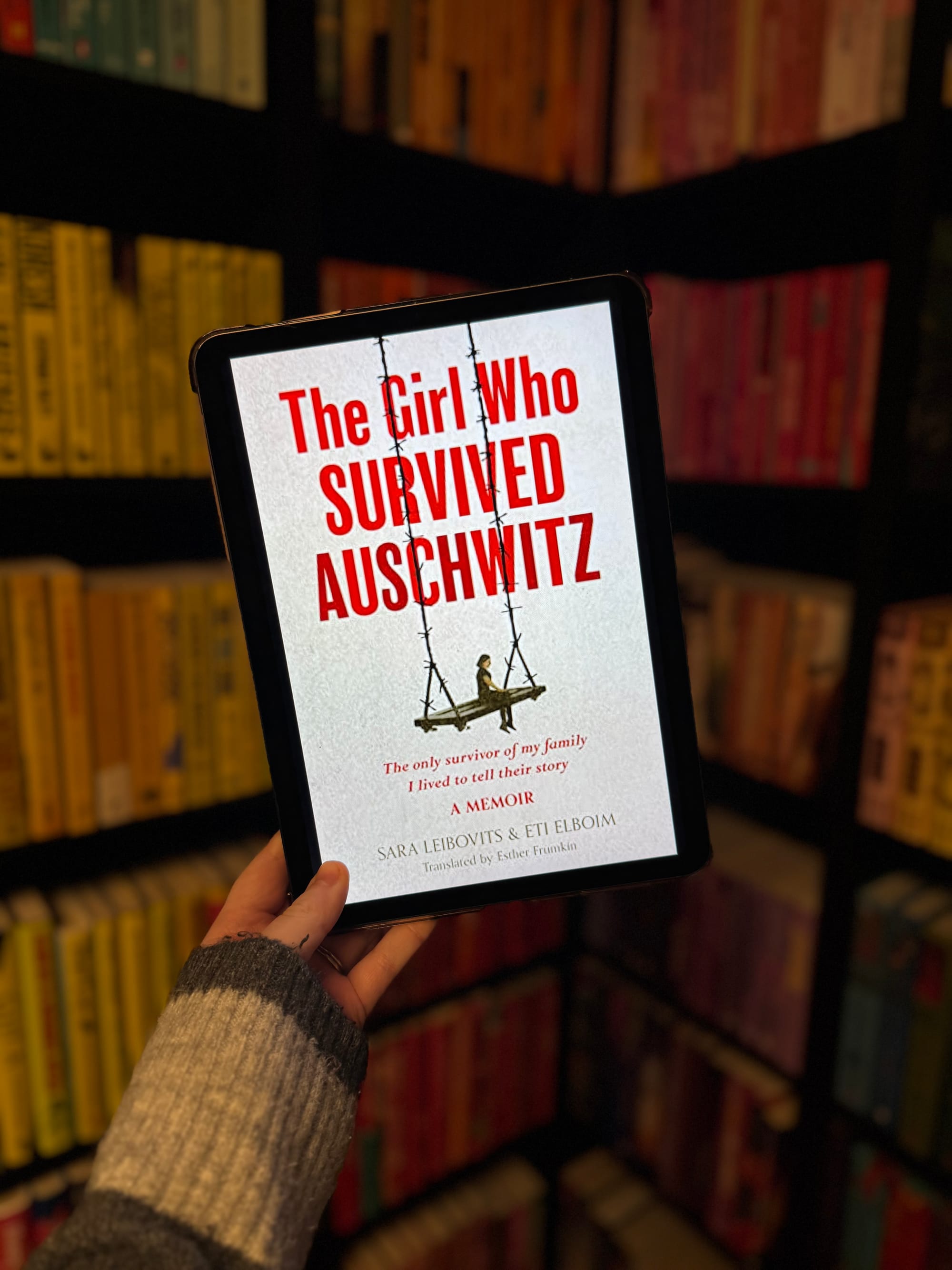 A hand holds a tablet displaying "The Girl Who Survived Auschwitz" book cover. Background shows bookshelves with colorful books.