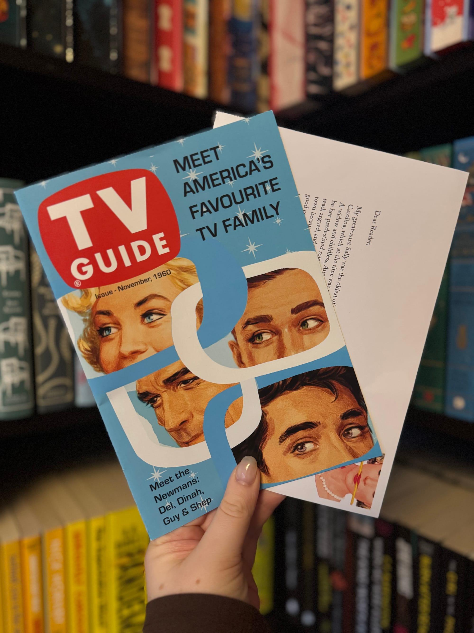 A hand holds a vintage style bookmark designed like a 1960 TV Guide cover reading “Meet America’s Favourite TV Family,” alongside an author letter. The bookmark shows four illustrated faces in mid century style, with a blurred bookshelf in the background.