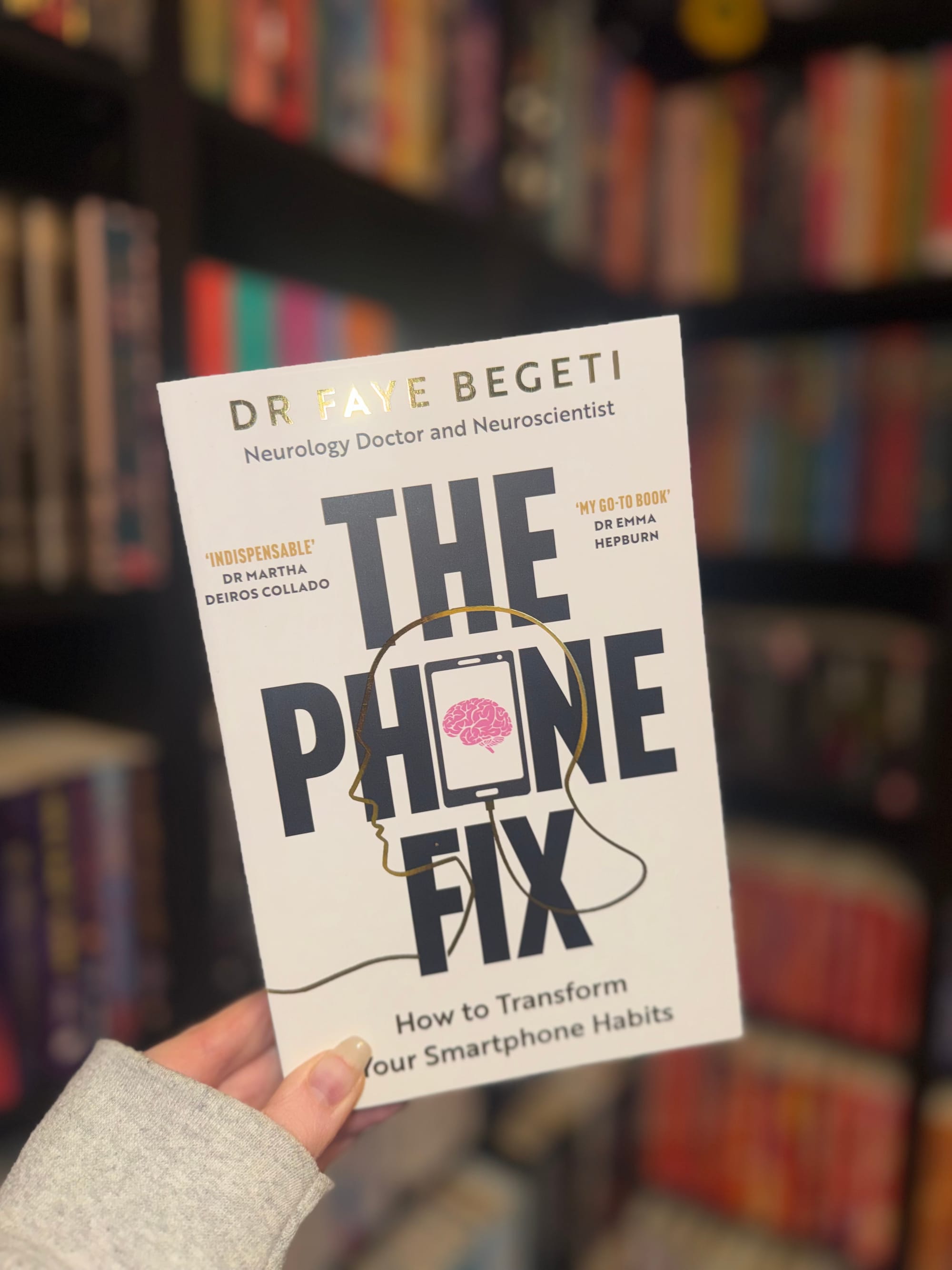 Hand holding a book titled "The Phone Fix" by Dr. Faye Begeti against a blurred colorful bookshelf background.