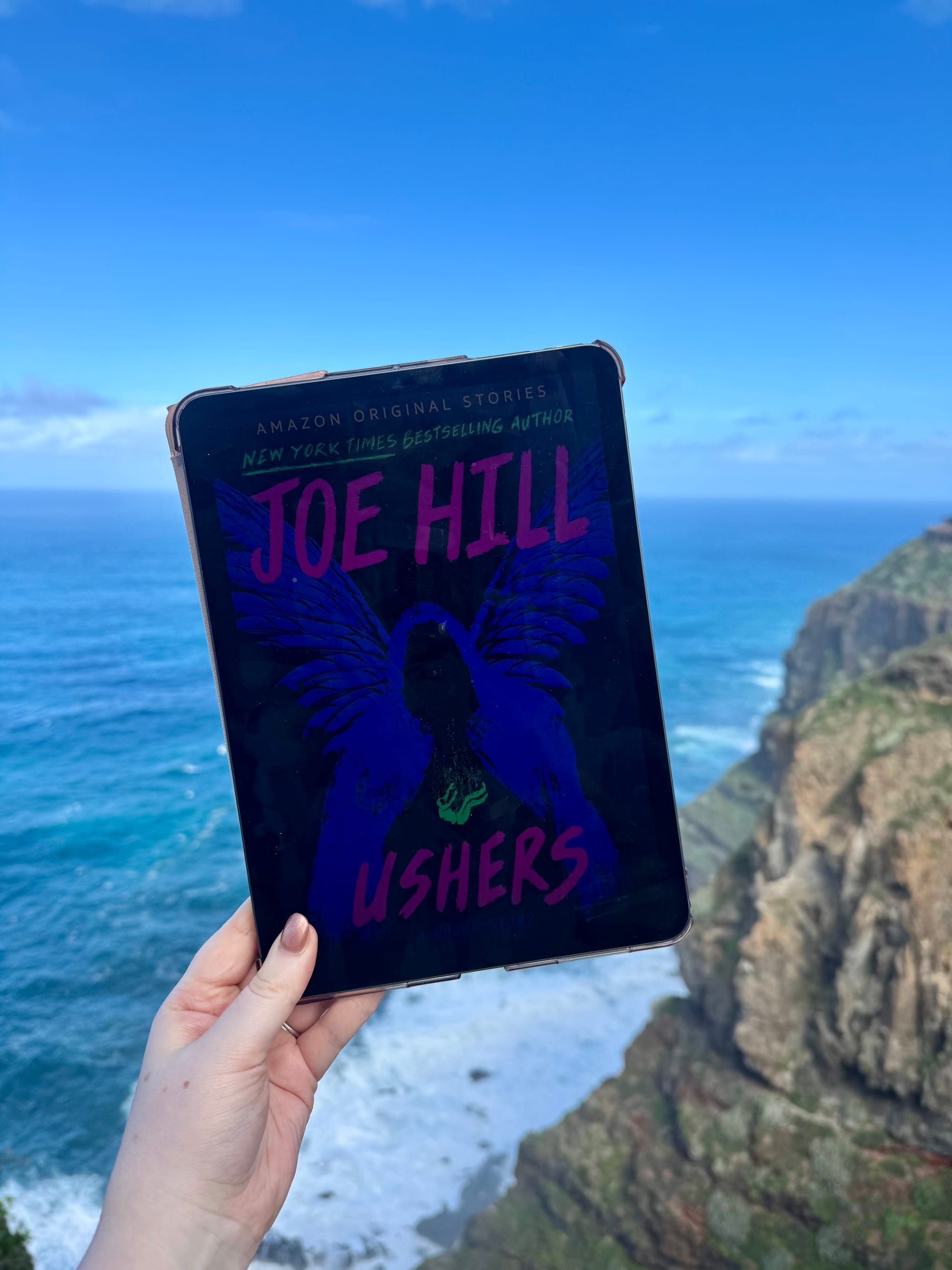 Hand holding a tablet displaying a book cover titled "Ushers" by Joe Hill. Background shows a rocky coastline and blue ocean under a clear sky.