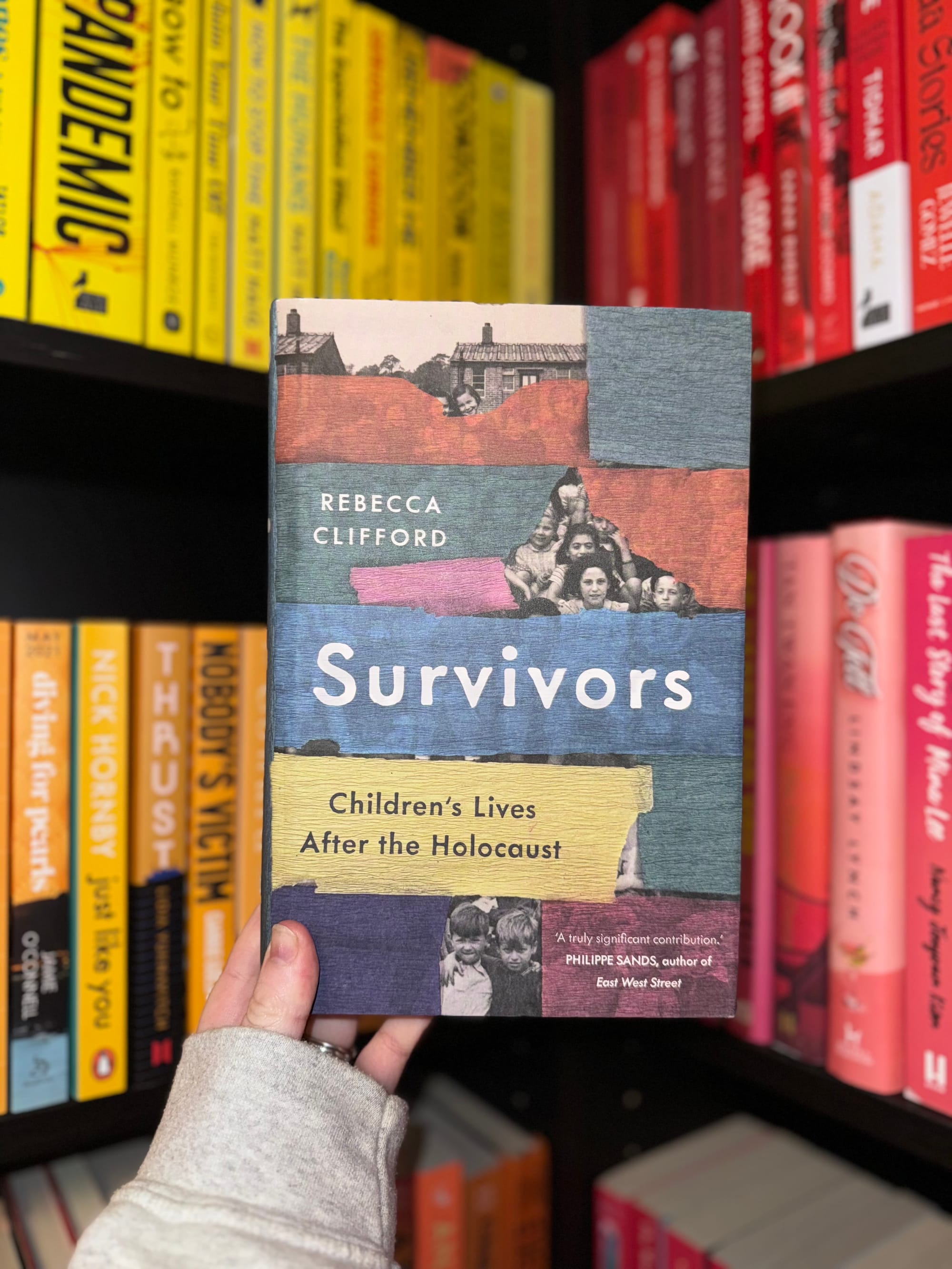 Hand holding "Survivors" book by Rebecca Clifford. Bookshelf background with colorful spines. Mood: historical and contemplative.