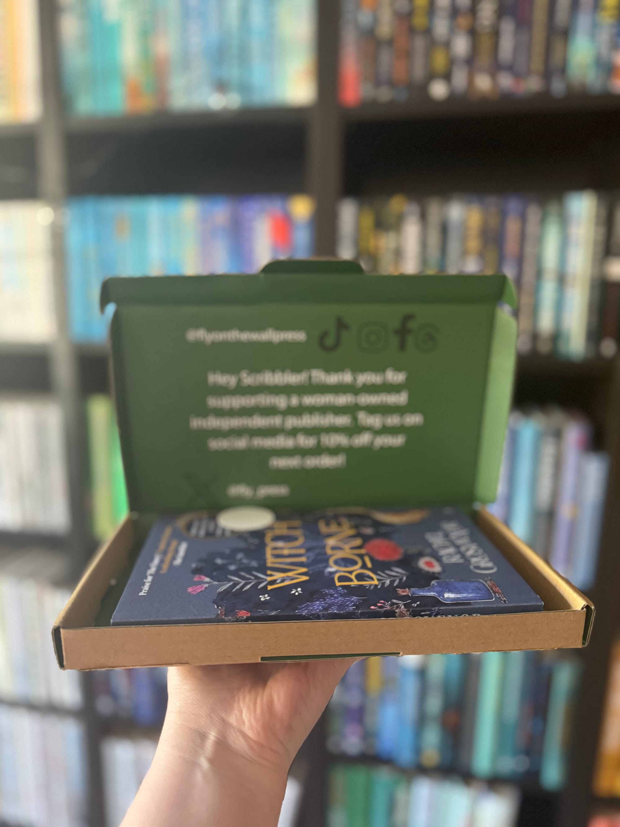Hand holding an open green box with a book titled "Witch of Bone" inside. Bookshelves with colorful books are blurred in the background. Enclosed text gives thanks for supporting a woman-owned publisher.