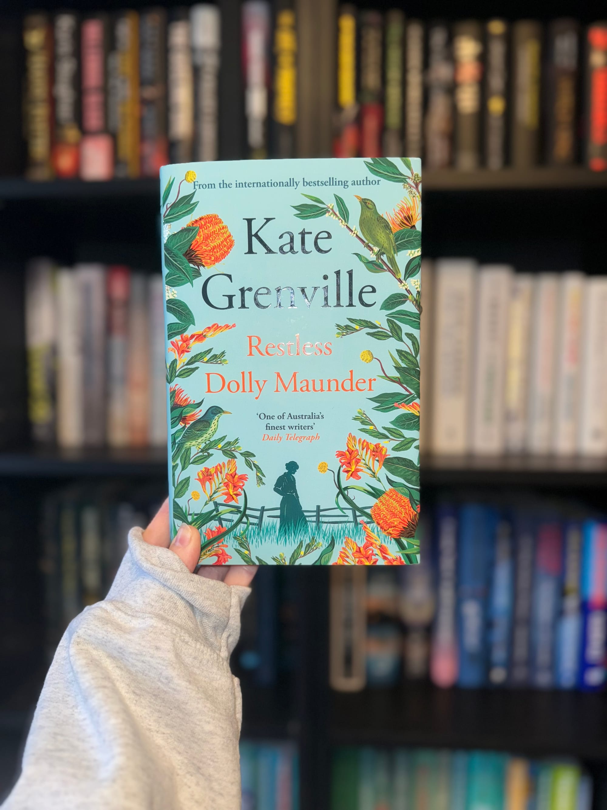 A hand holds a book titled "Restless Dolly Maunder" by Kate Grenville, with a floral cover design. Blurred bookshelves in the background.