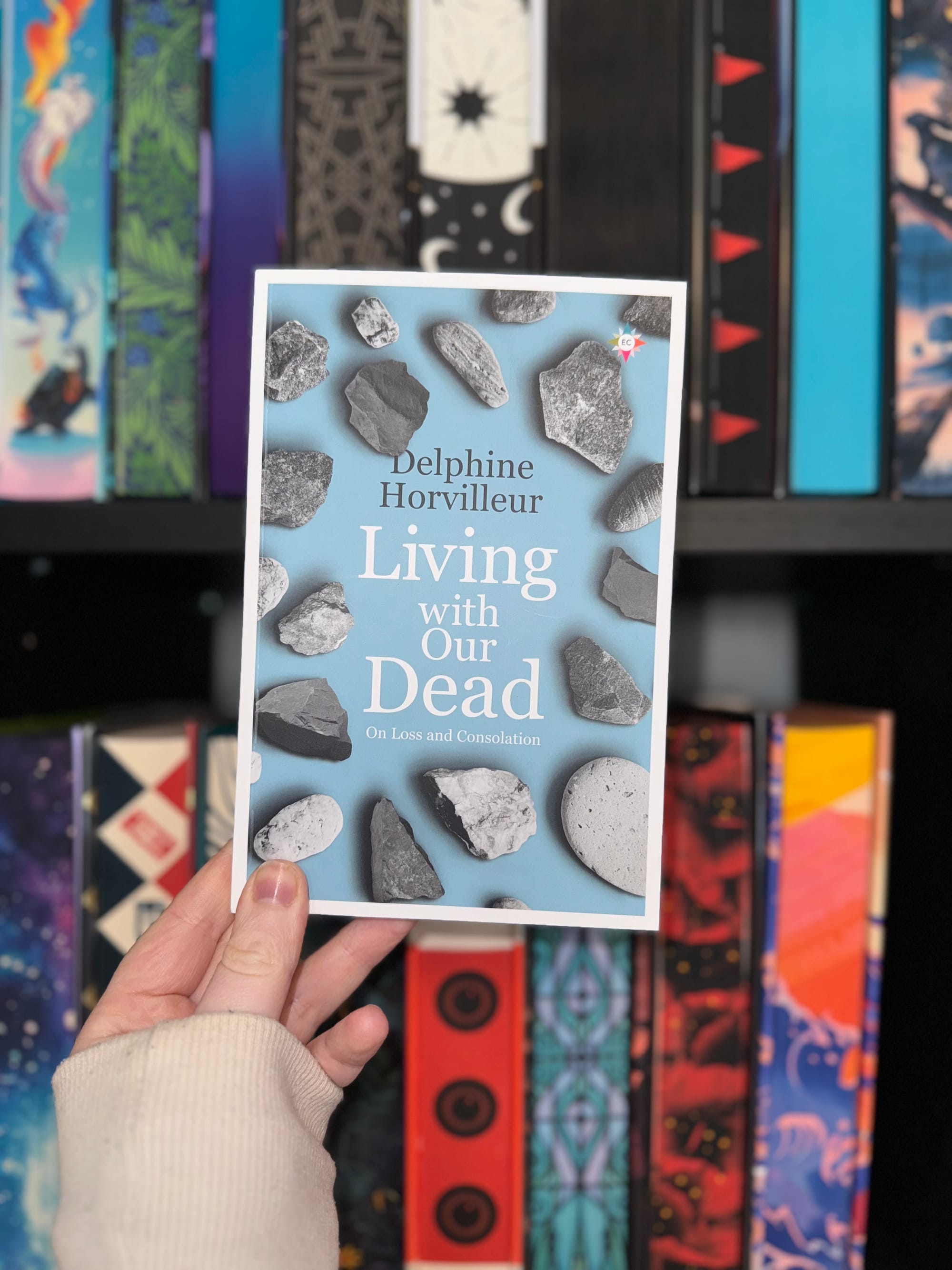 Hand holding a book titled "Living with Our Dead" by Delphine Horvilleur. Background shows colorful patterned books on a shelf.
