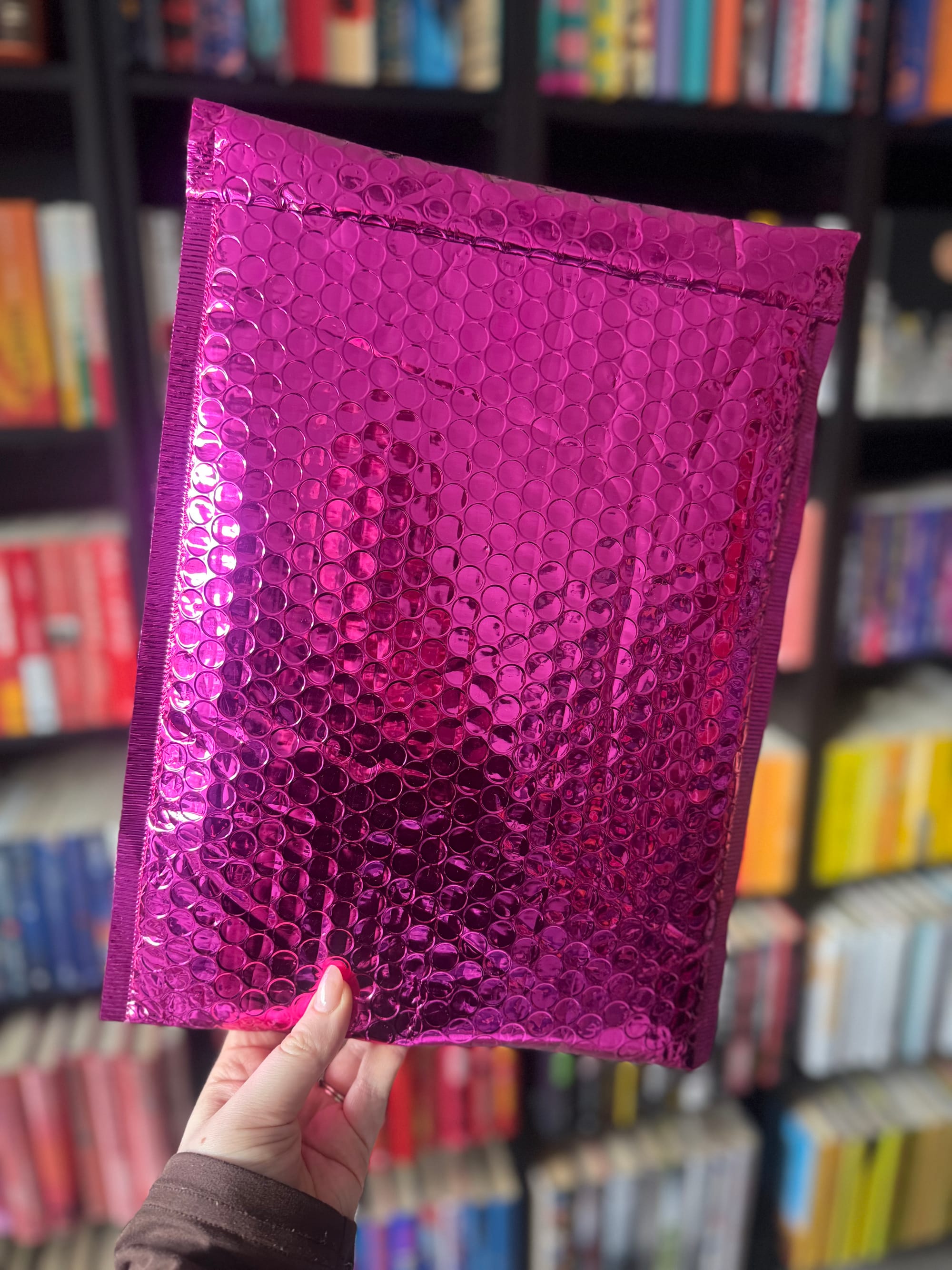 A hand holding a bright metallic pink padded envelope in front of a blurred bookshelf filled with colourful books.