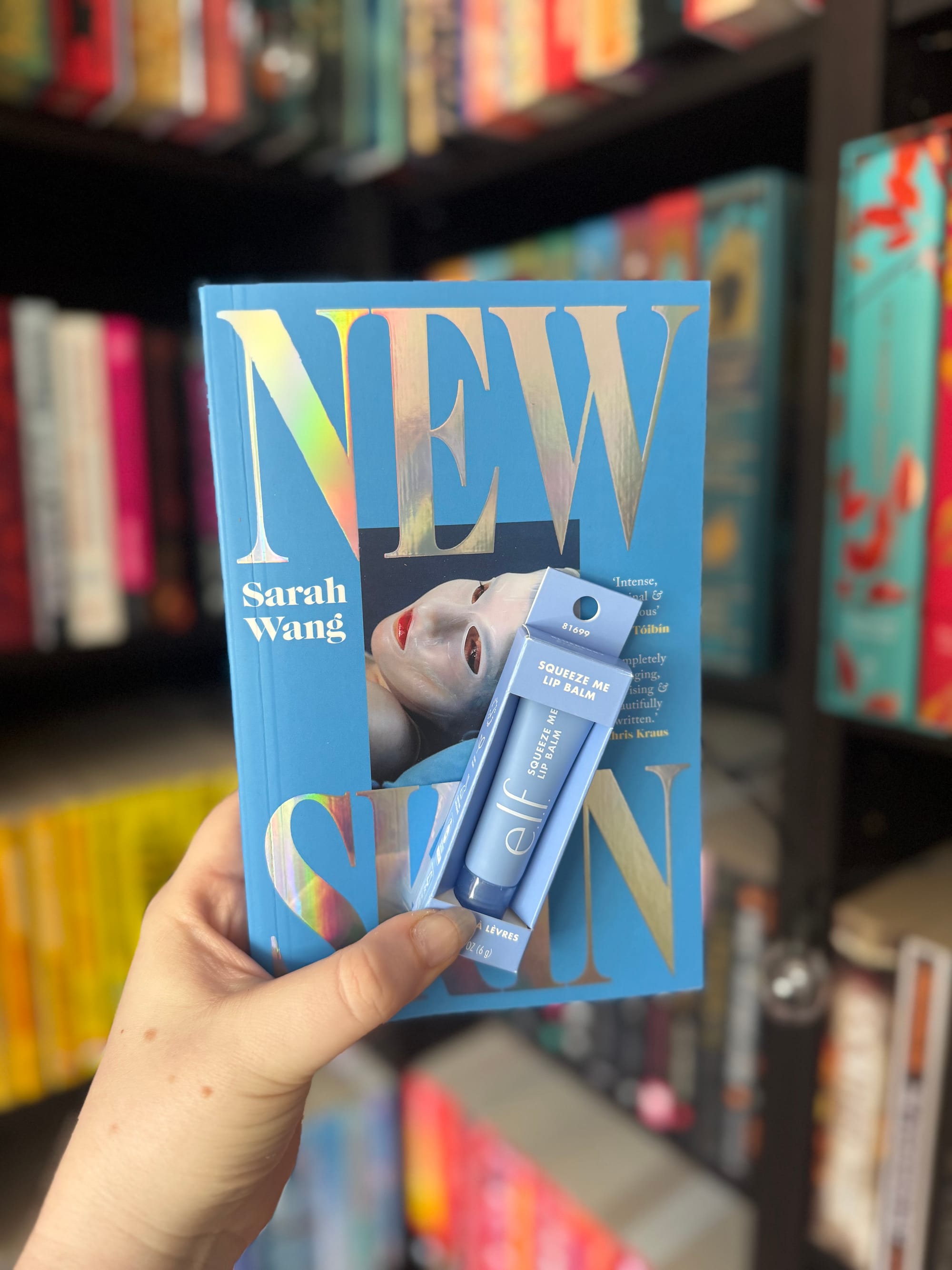 Hand holding a blue book titled "NEW SKIN" by Sarah Wang, alongside a blue tube of elf lip balm, with colorful bookshelves in the background.