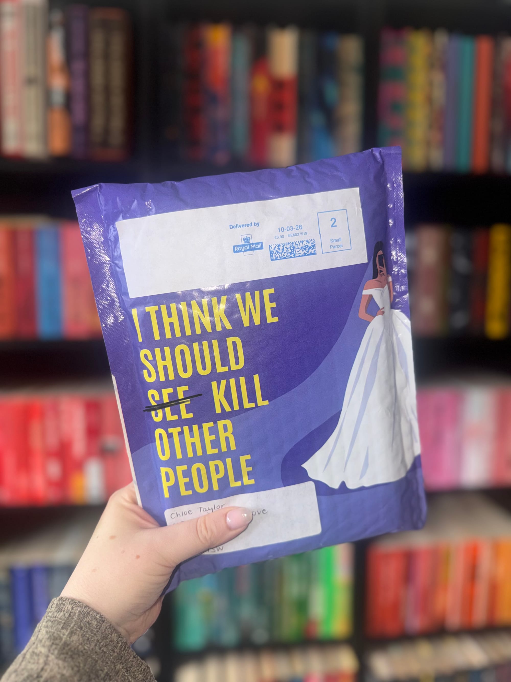 A hand holds a purple package with yellow text saying "I THINK WE SHOULD KILL OTHER PEOPLE." Bookshelves with colorful books are in the background.