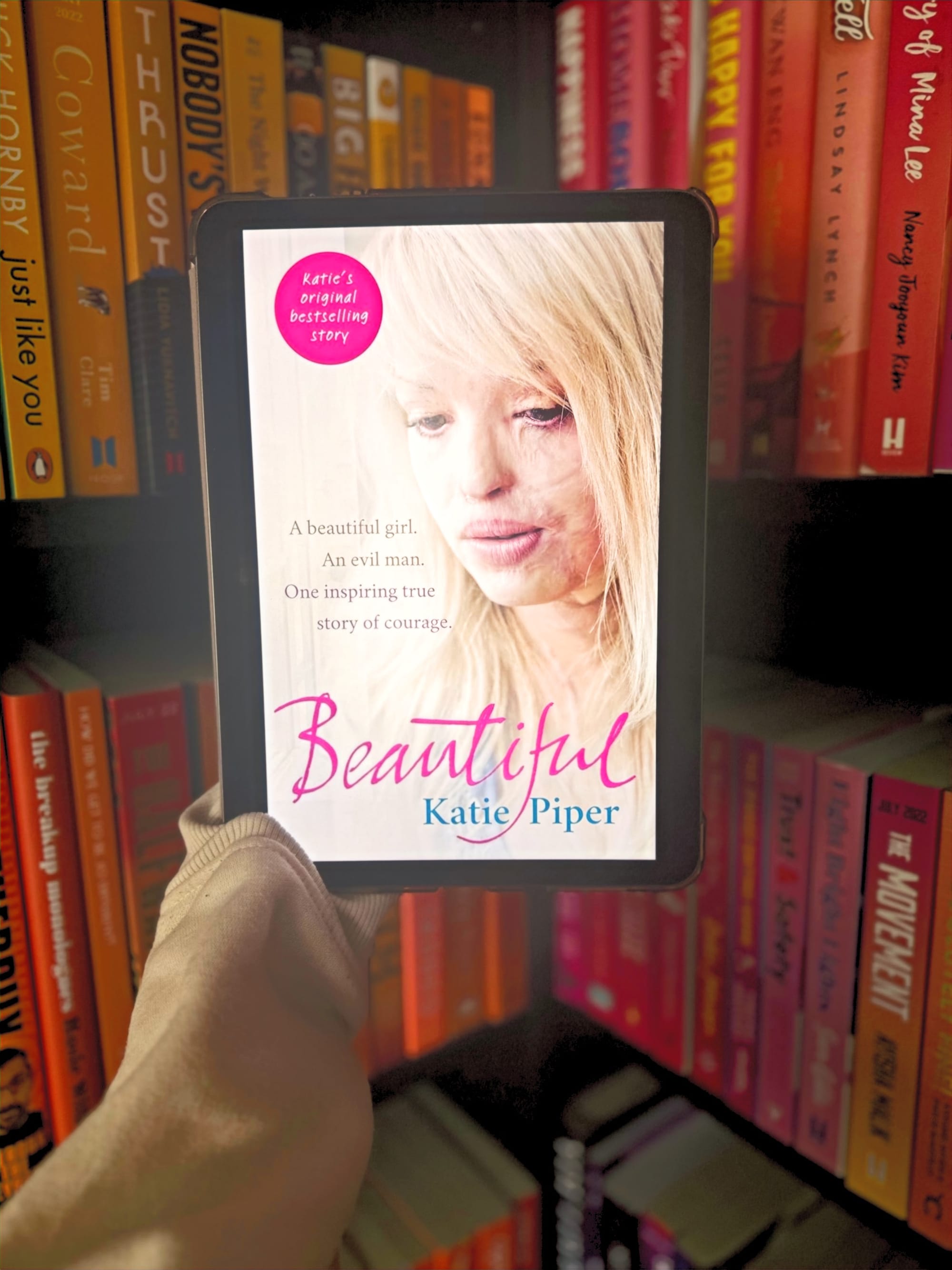 Tablet displaying "Beautiful" by Katie Piper held before vibrant bookshelf. Pink text highlights an inspiring true story of courage.
