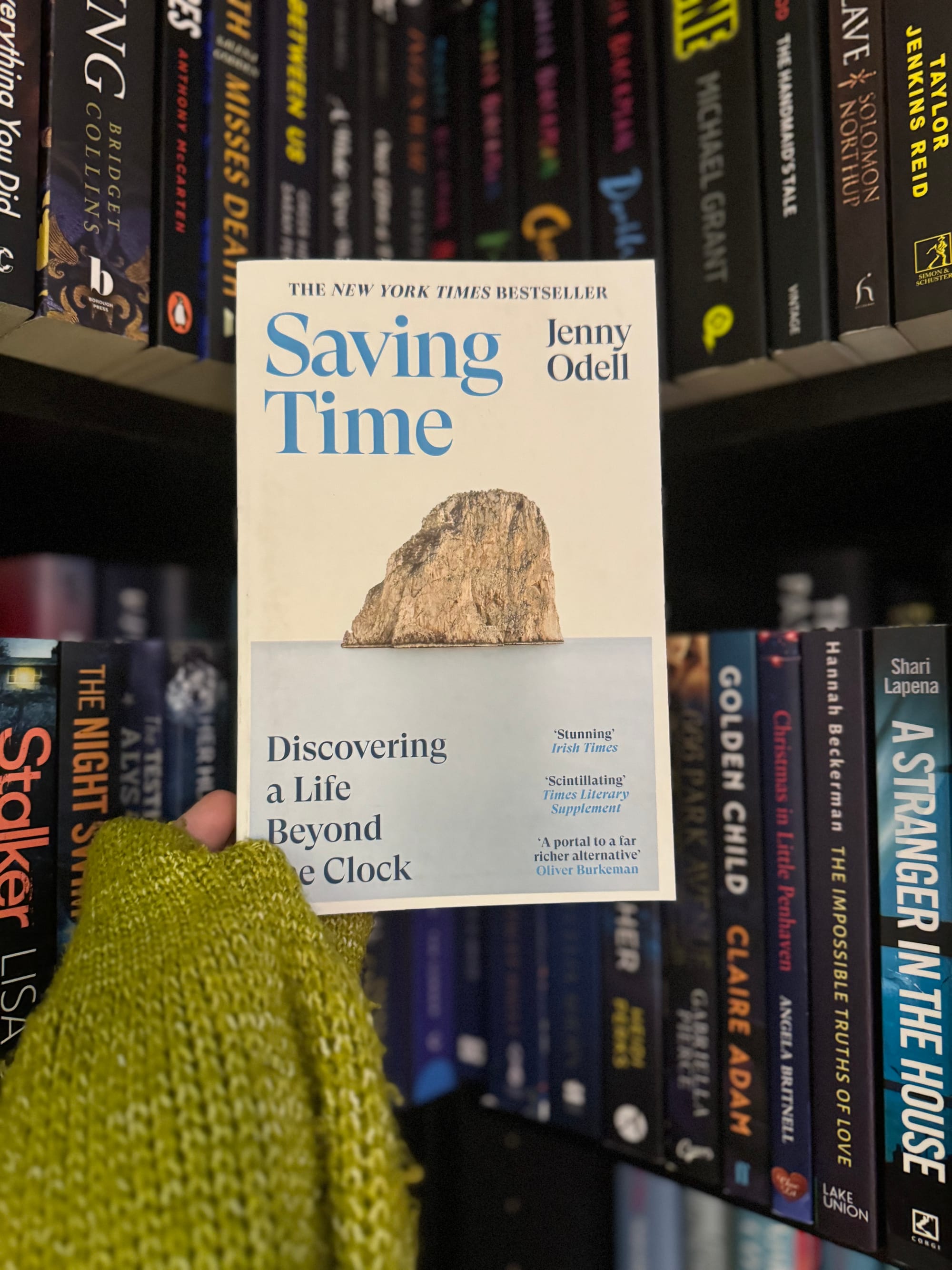 Hand in green sweater holds the book "Saving Time" by Jenny Odell against a backdrop of colorful books on shelves.