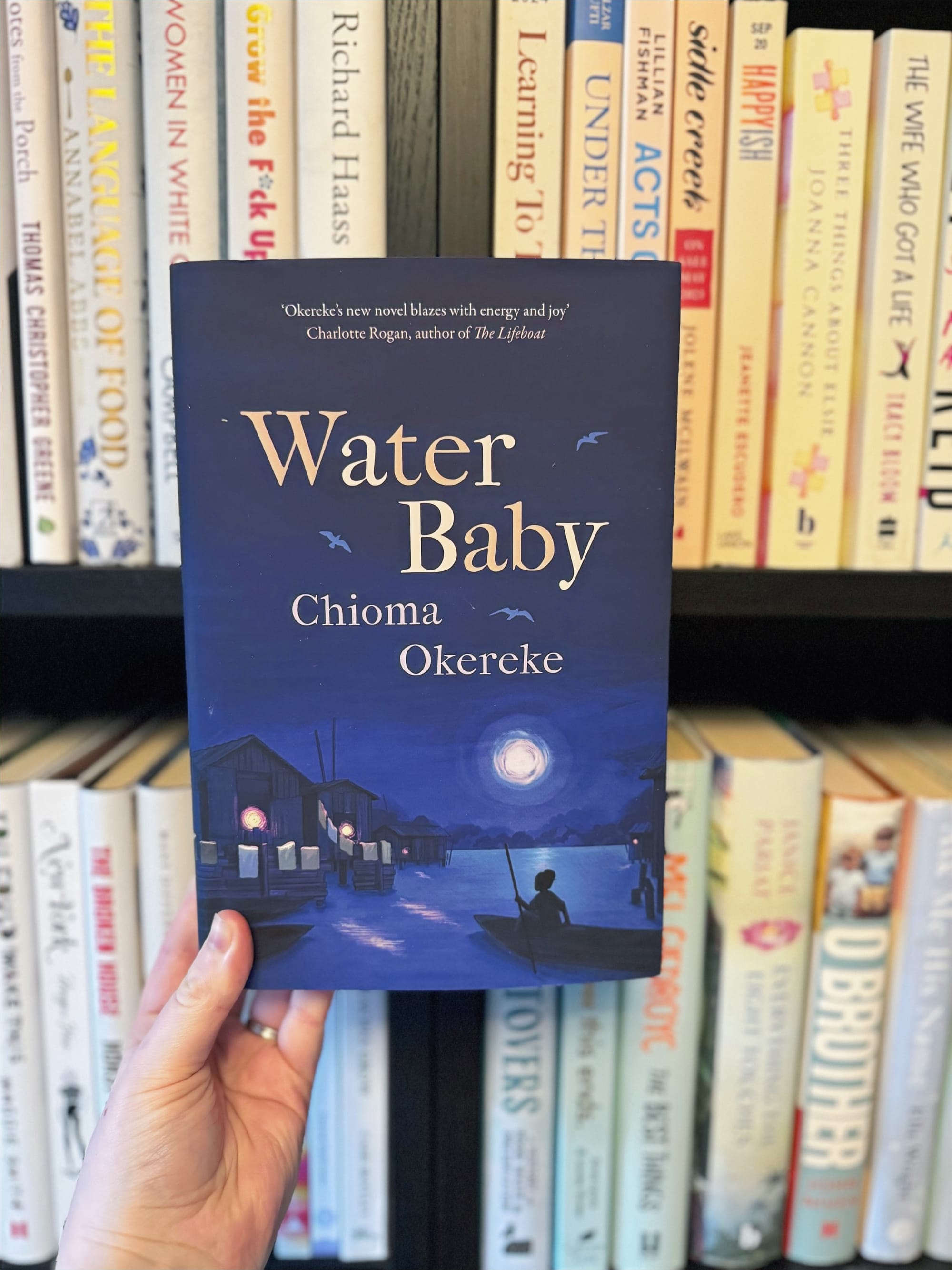 A hand holds the book "Water Baby" by Chioma Okereke against a bookshelf. The cover shows a night scene with huts, water, and a boat.