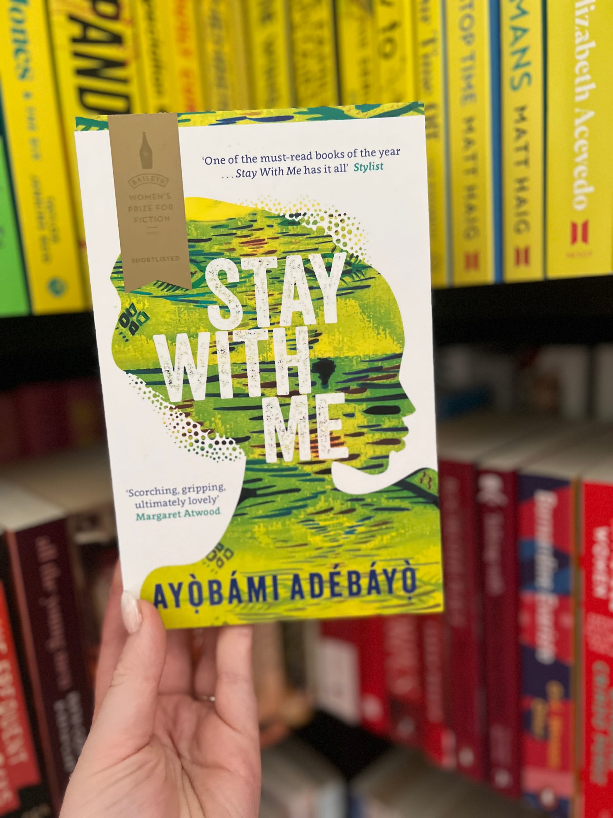 A hand holds the book "Stay With Me" by Ayọ̀bámi Adébáyọ̀ against colorful bookshelves. The cover shows text and abstract green-yellow art.