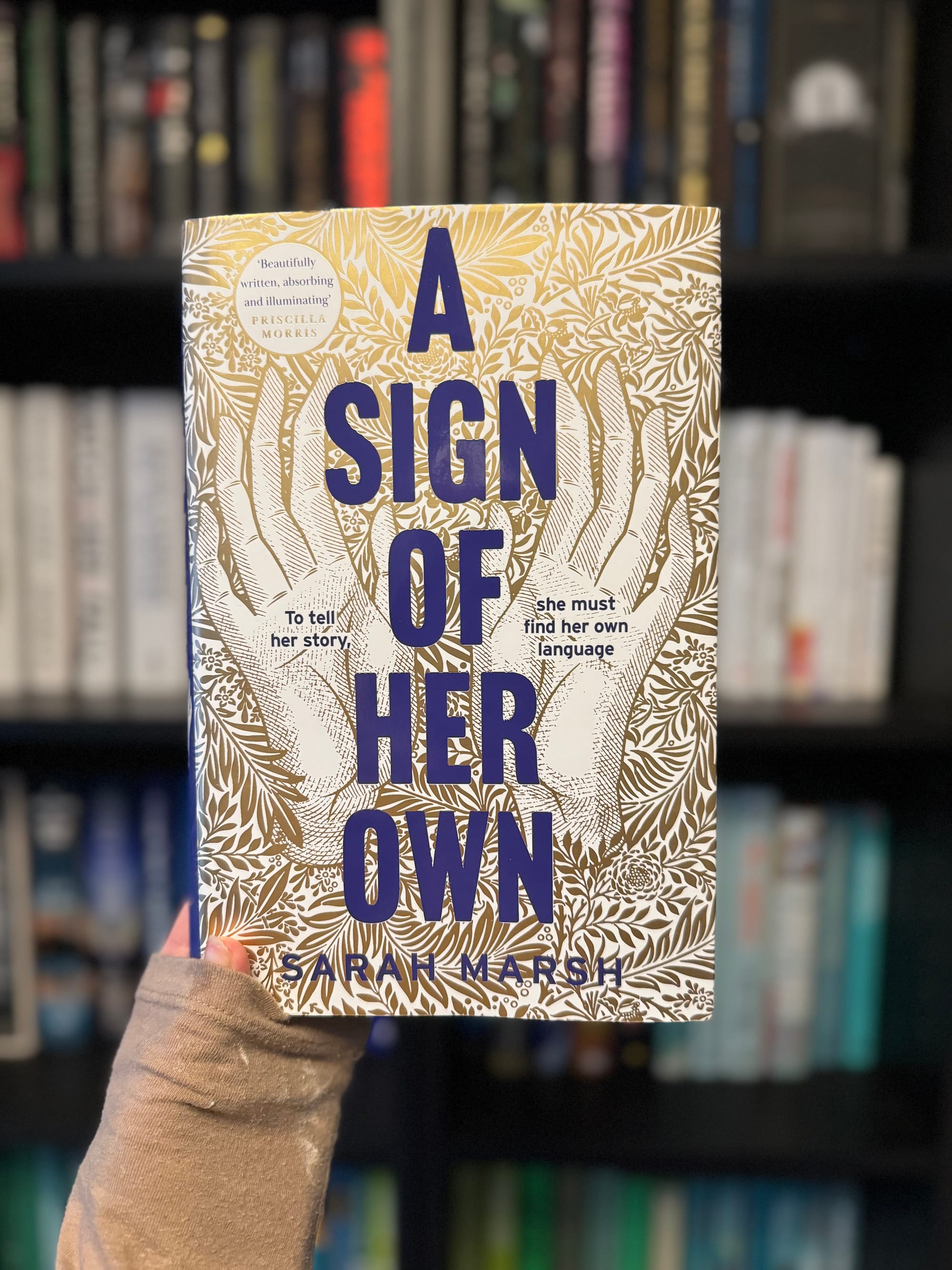 A hand holds "A Sign of Her Own" book with blue text and gold leaf pattern on cover, set against a blurred bookshelf background.