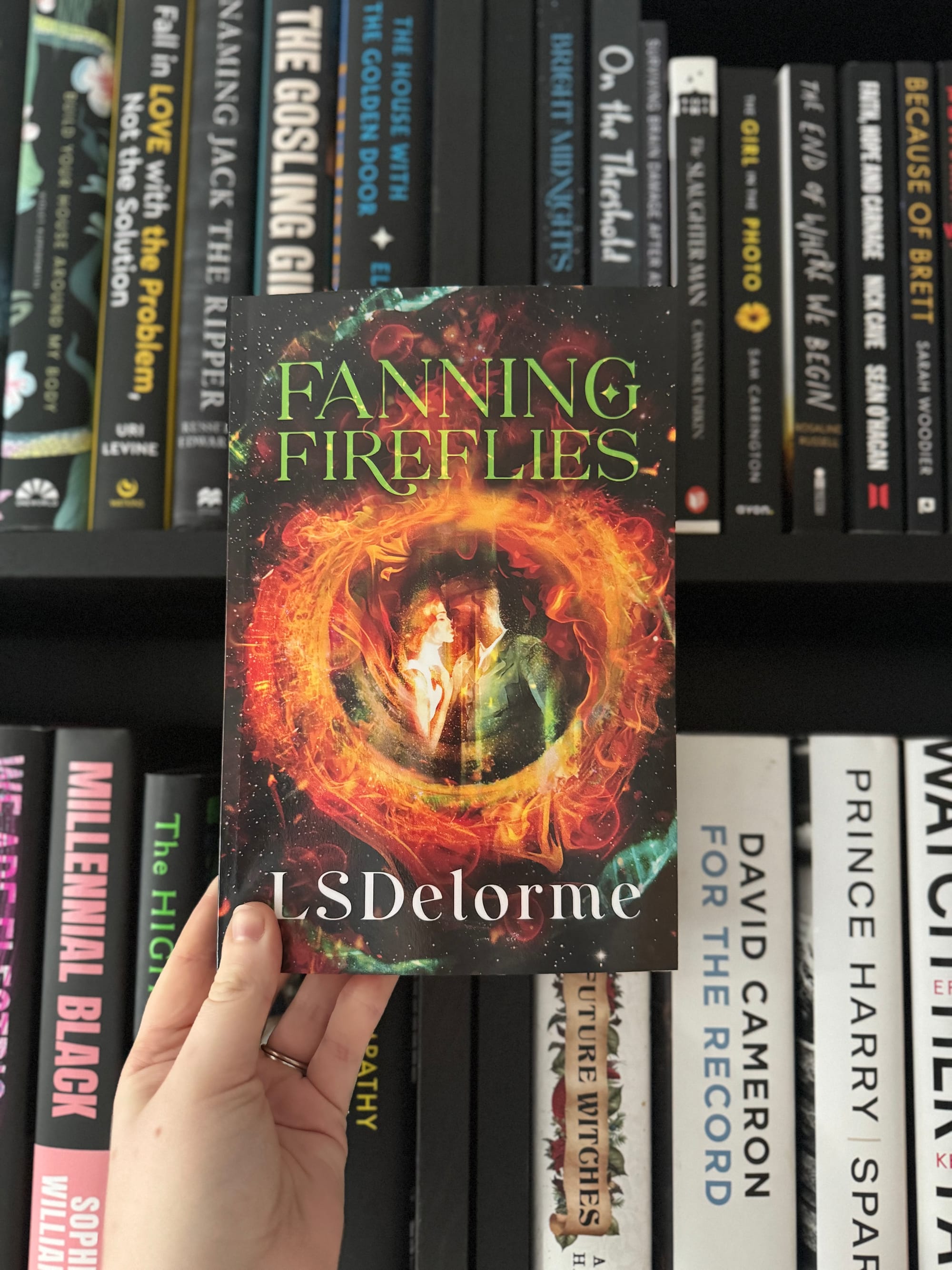 A person holds a book titled "Fanning Fireflies" by LS Delorme in front of a bookshelf. The cover features fiery and mystical artwork.