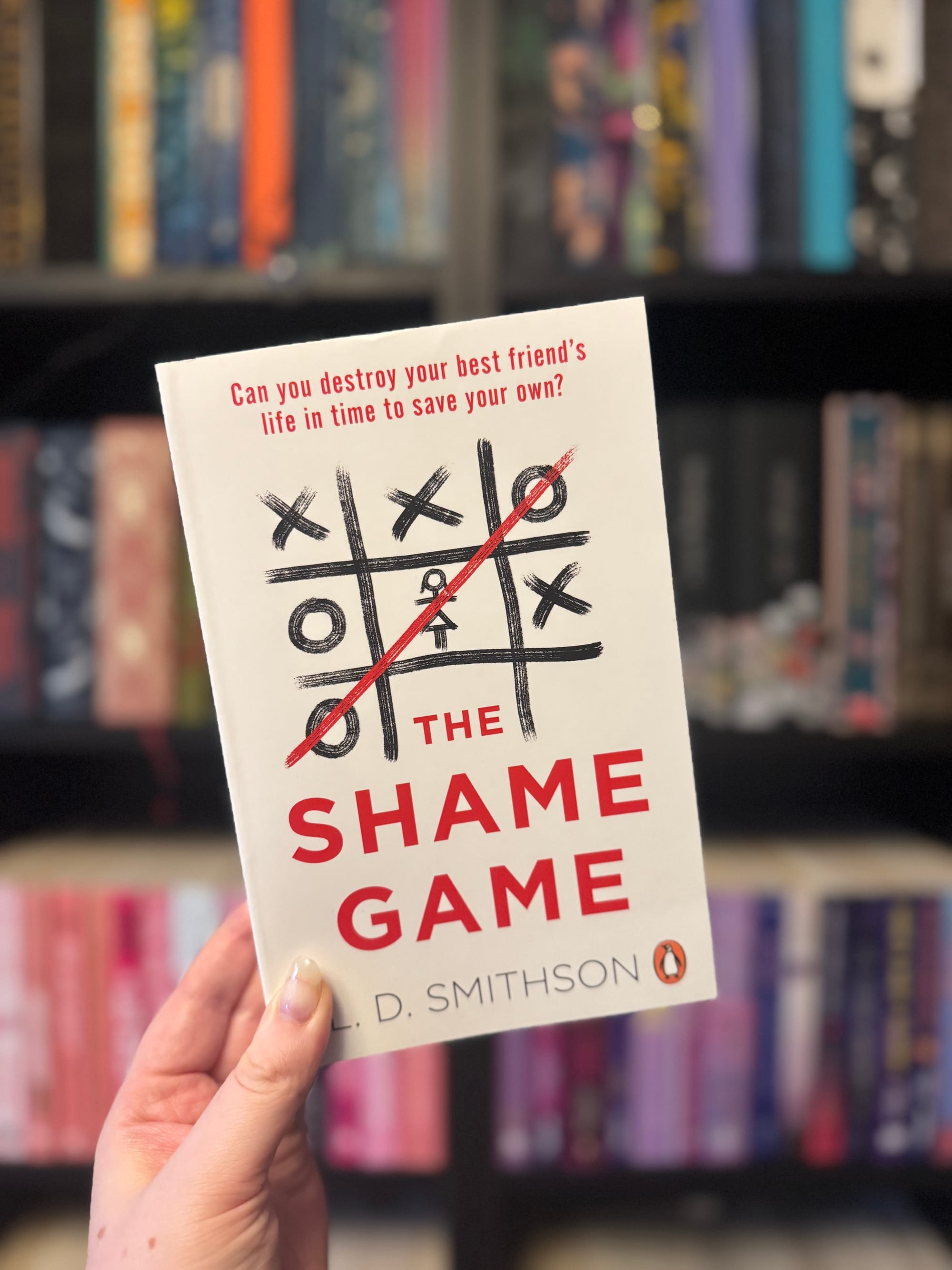Hand holding a book titled "The Shame Game" with a tic-tac-toe design. Bookshelf with colorful books in the blurred background.
