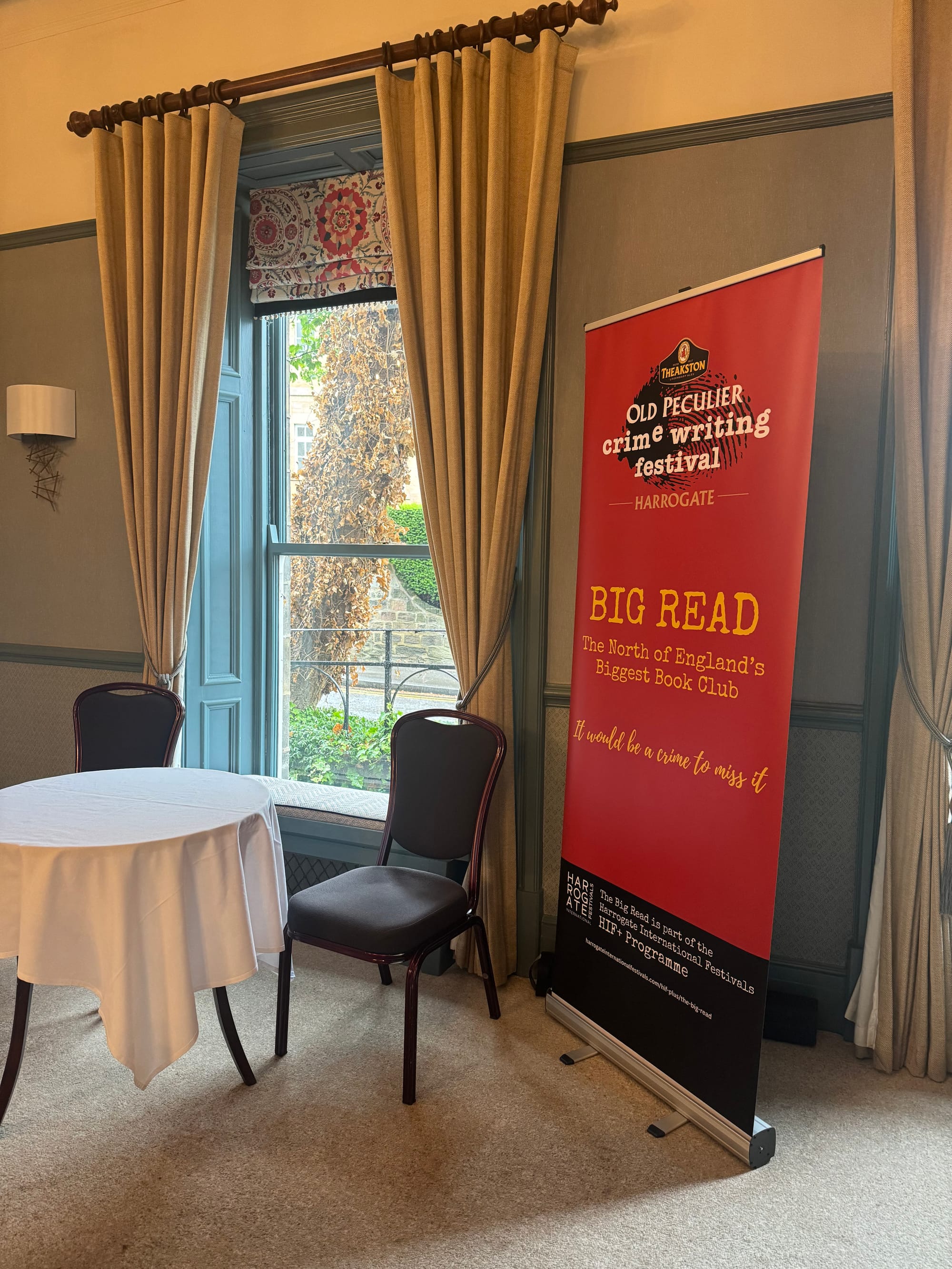 Room with a table, chairs, and a red banner for the "Old Peculier Crime Writing Festival" by a large window with beige curtains.