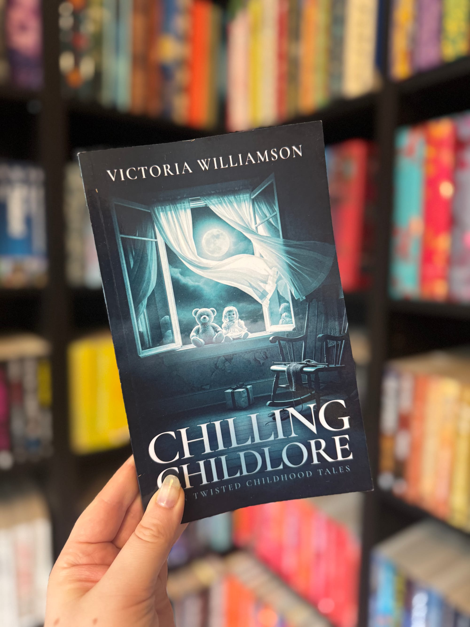 A hand holds the book Chilling Childlore: Twisted Childhood Tales by Victoria Williamson, showing a dark blue cover with two teddy bears sitting at a window under the moonlight, with shelves of bright books blurred in the background.