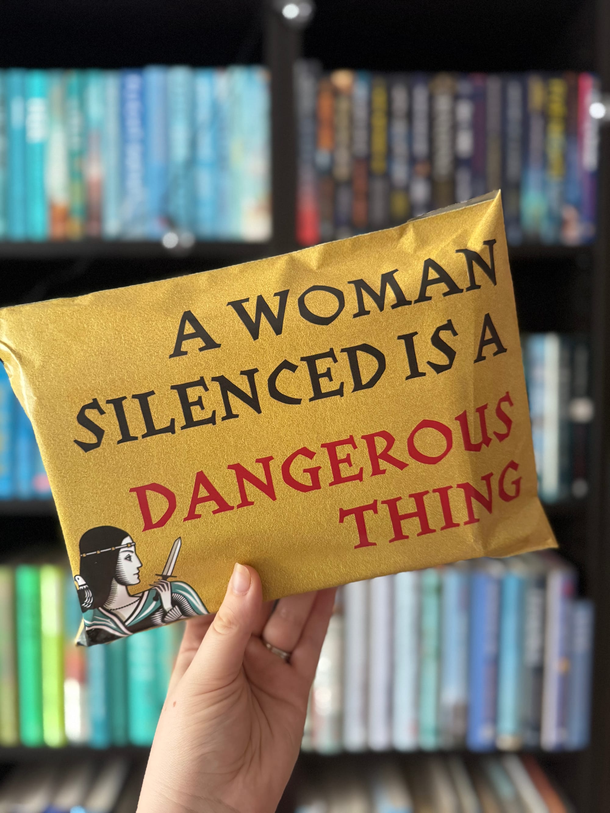 Hand holds gold package with "A Woman Silenced is a Dangerous Thing" text in black and red. Bookshelves with colorful spines in background.
