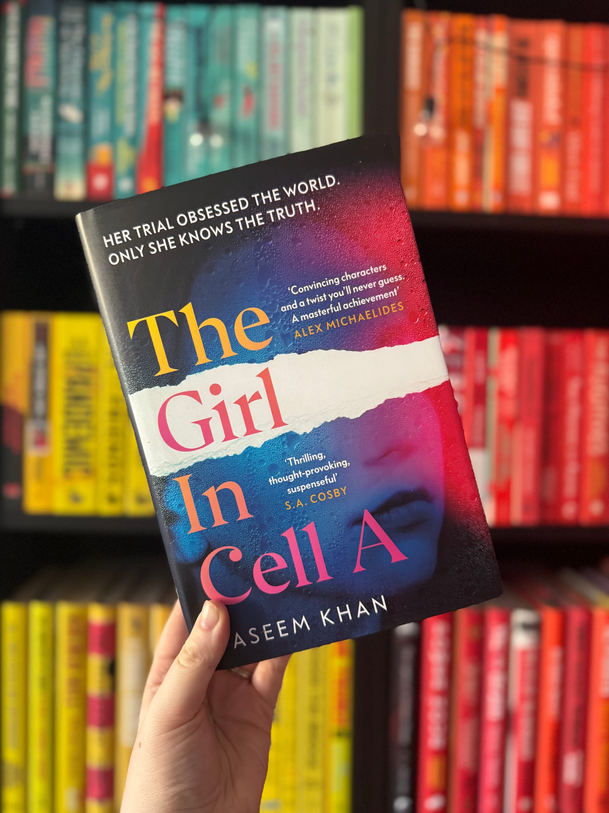A hand holds the book "The Girl In Cell A" against a colorful bookshelf. The cover features a woman's face and bold text blurbs.