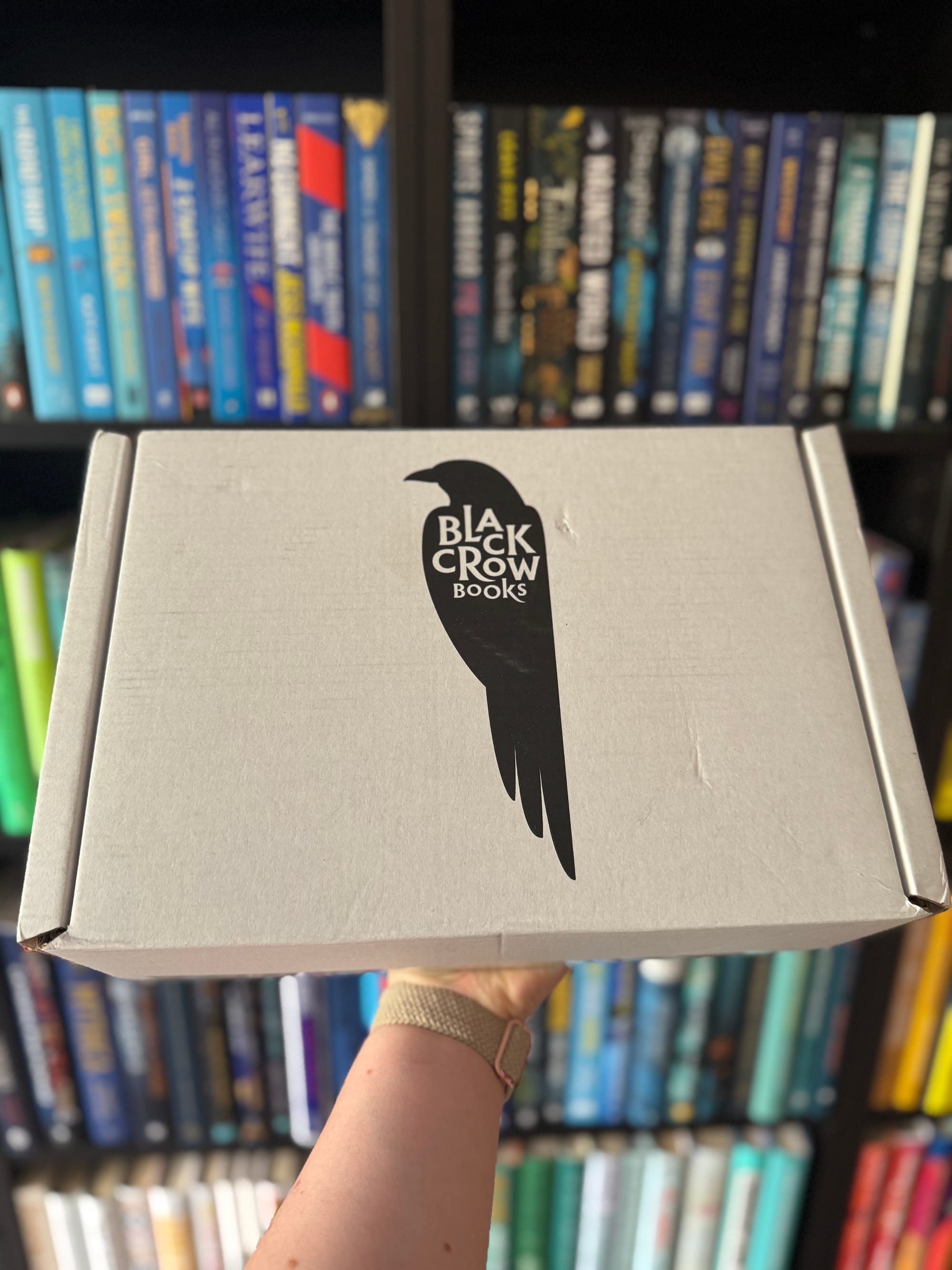 A hand holding up a grey cardboard box with a black silhouette of a crow and the text 'Black Crow Books' printed on it. In the background, there are shelves filled with colourful books arranged by spine colour.