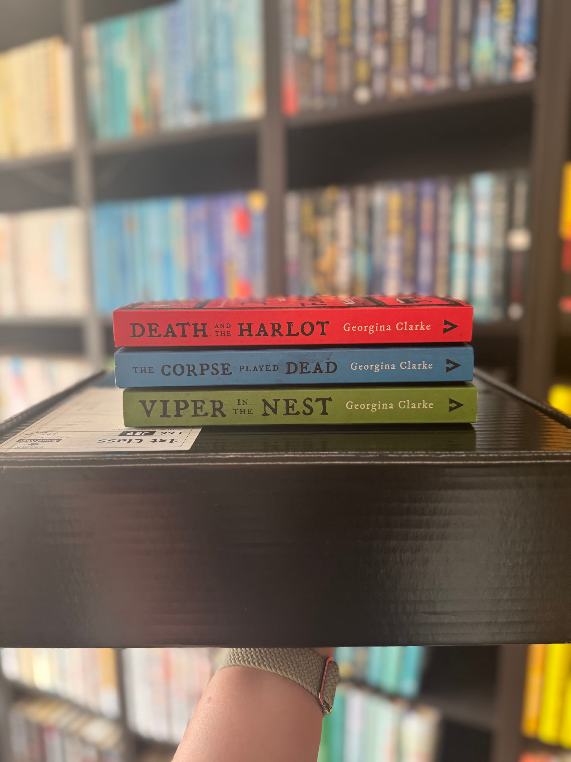 Stack of three colorful books by Georgina Clarke on a black shelf in a bookstore. A hand with a wristband holds the shelf.