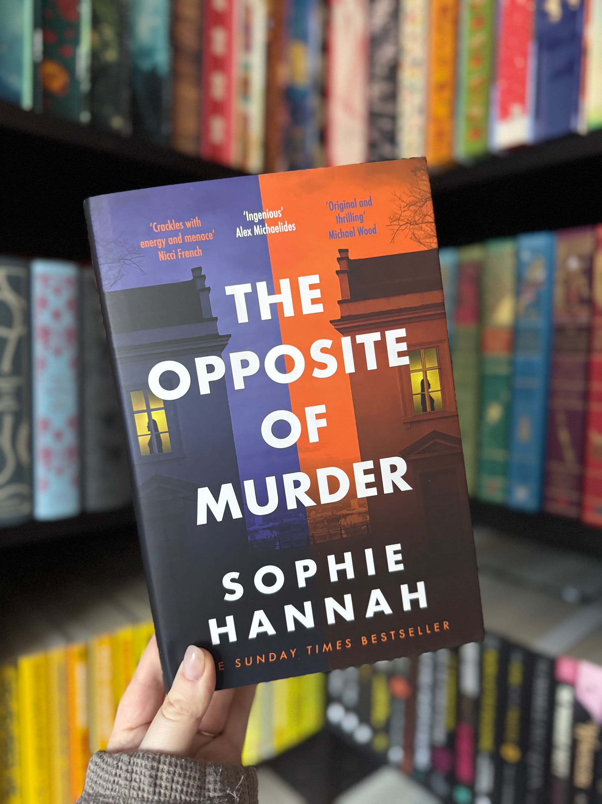 A hand holds "The Opposite of Murder" by Sophie Hannah, set against colorful bookshelves. Cover features blue and orange tones.