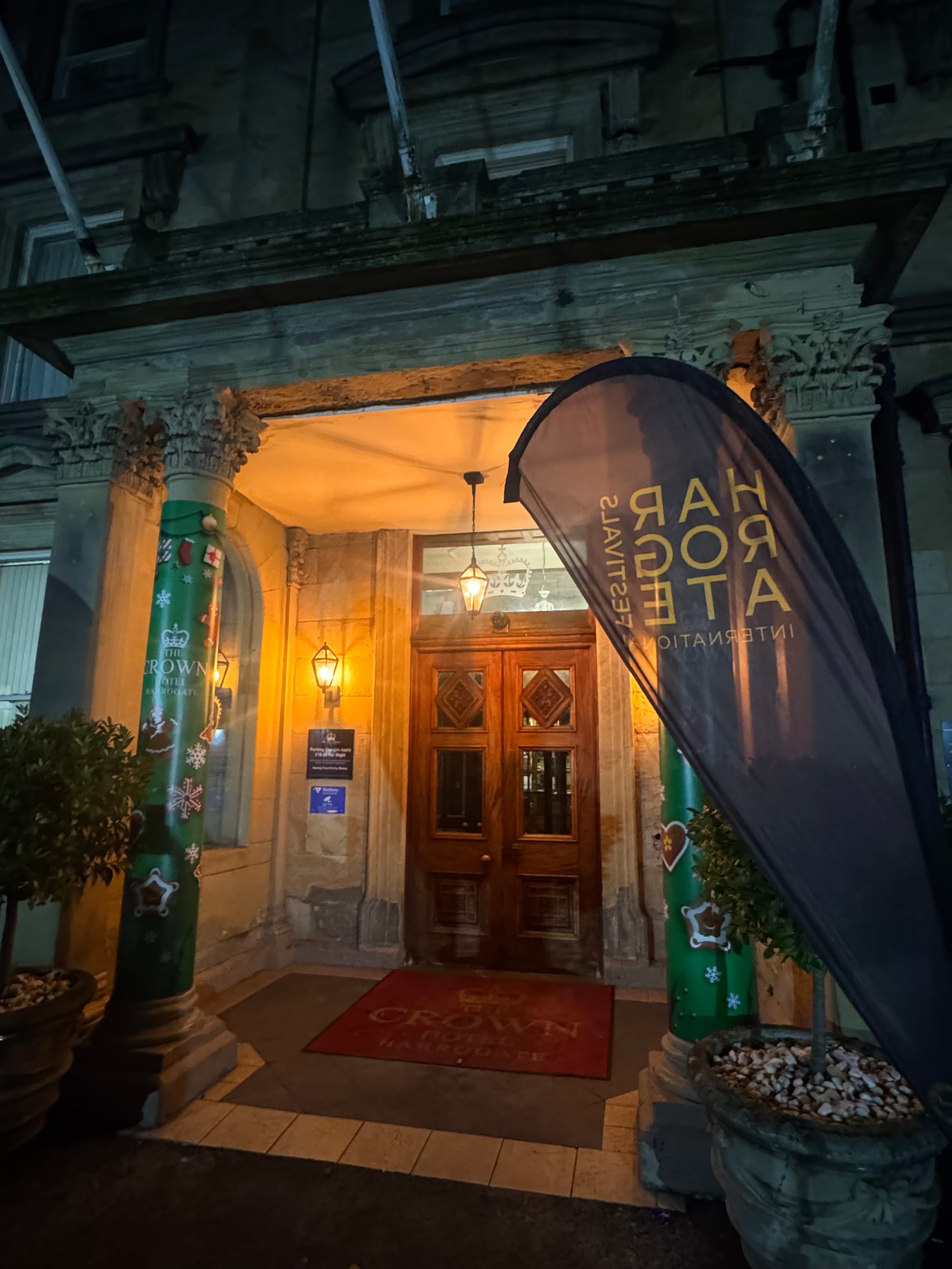 Nighttime hotel entrance with ornate columns, holiday decorations, and warm lighting. "Crown Hotel" text on red mat. Vertical banner nearby.