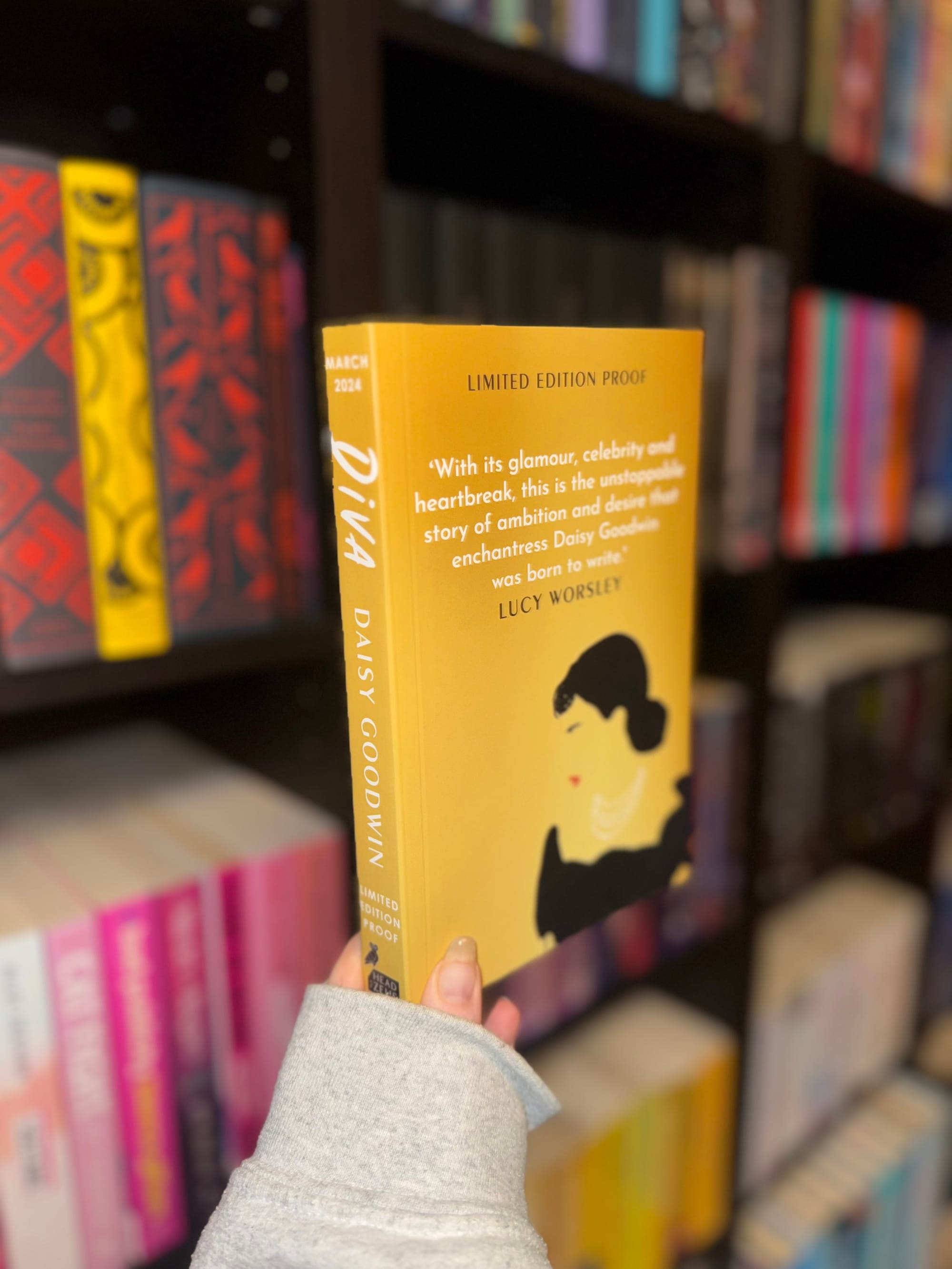 A person holds a yellow book titled Diva by Daisy Goodwin in front of a blurry bookshelf. The cover features a woman’s silhouette.