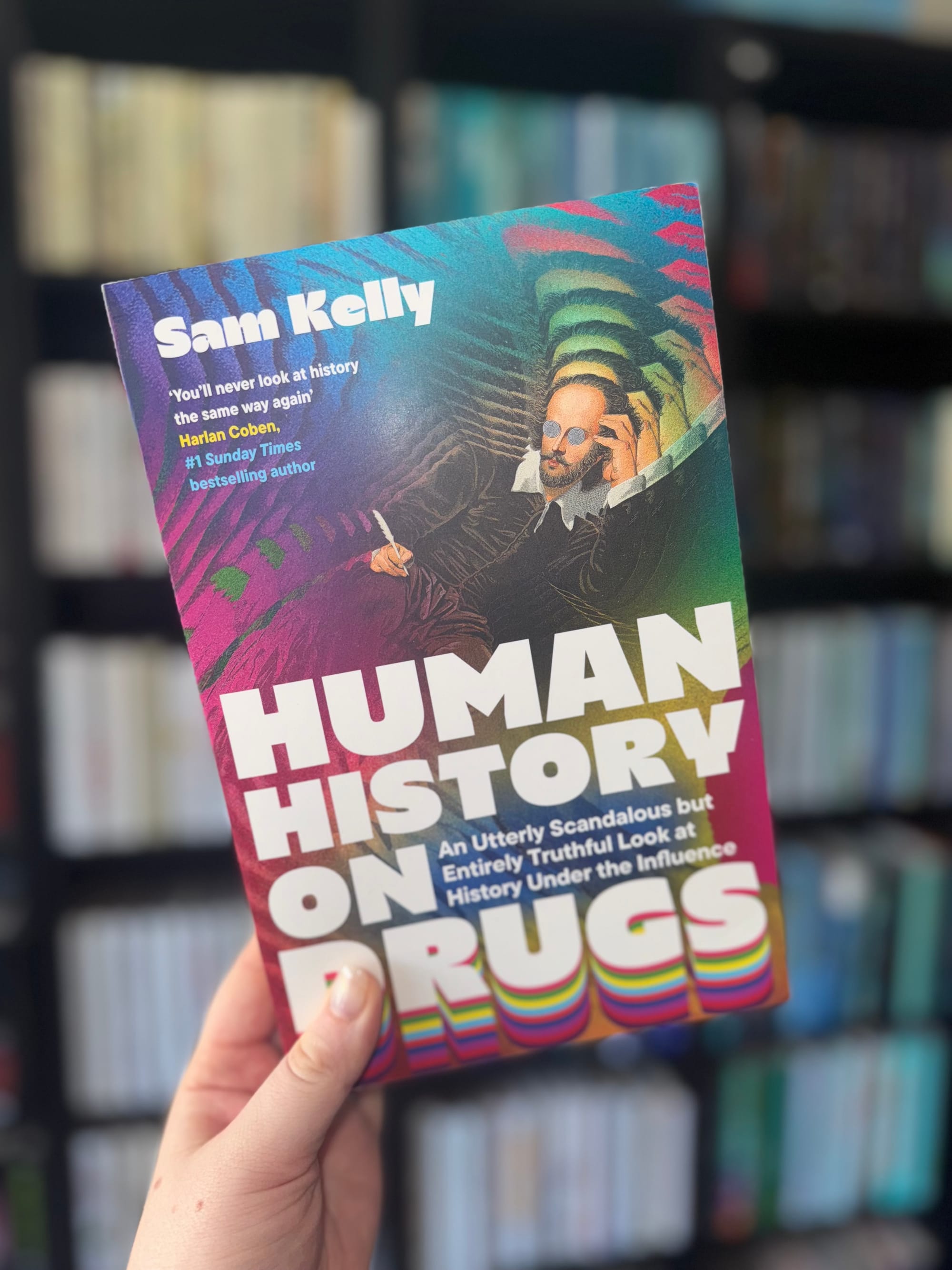 Hand holds a colorful book titled "Human History on Drugs" by Sam Kelly. Background shows blurred bookshelf. Text excerpt mentions history.