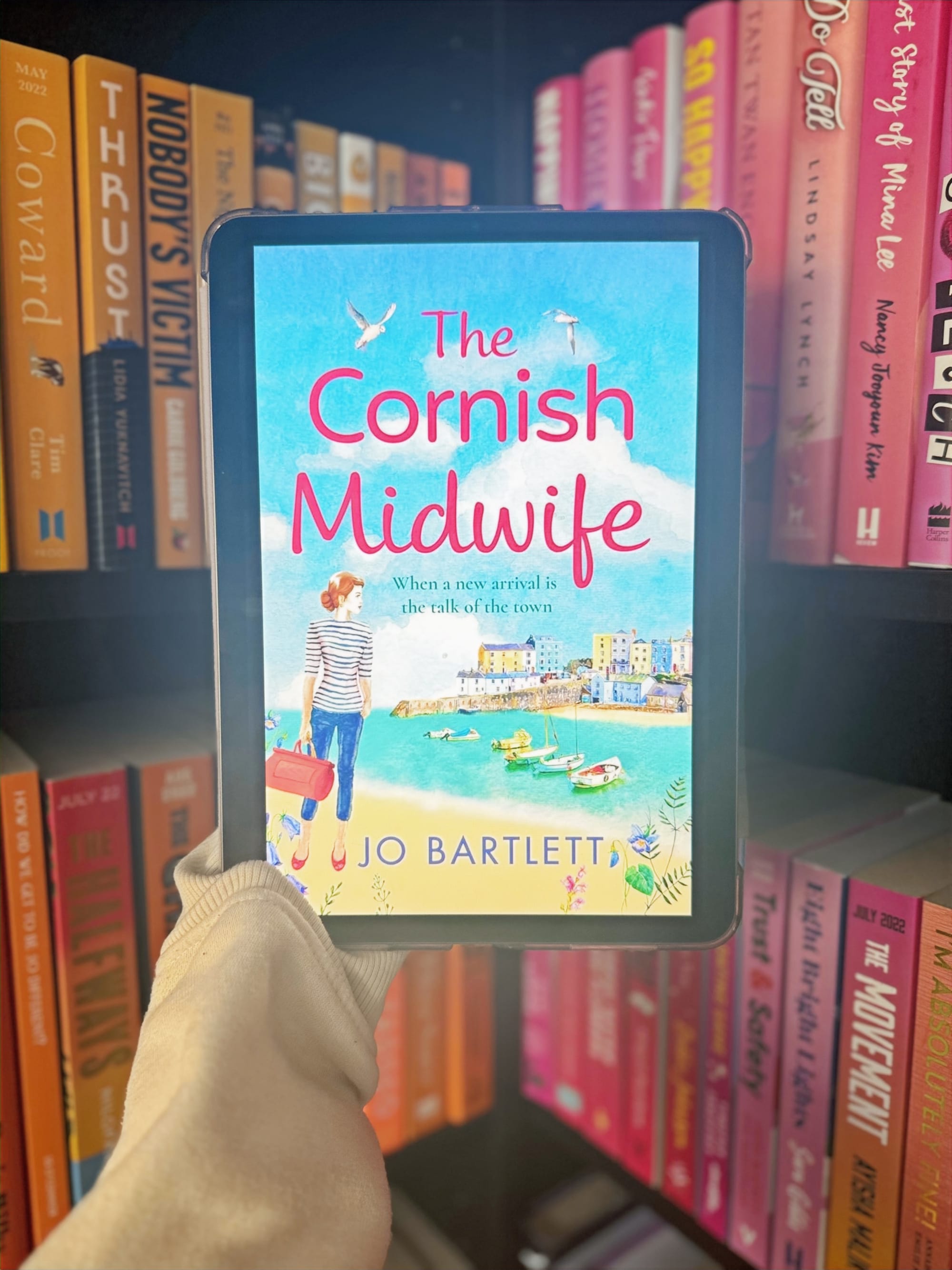 E-reader displaying "The Cornish Midwife" book cover in colorful book-filled library. Cover shows seaside scene with text by Jo Bartlett.