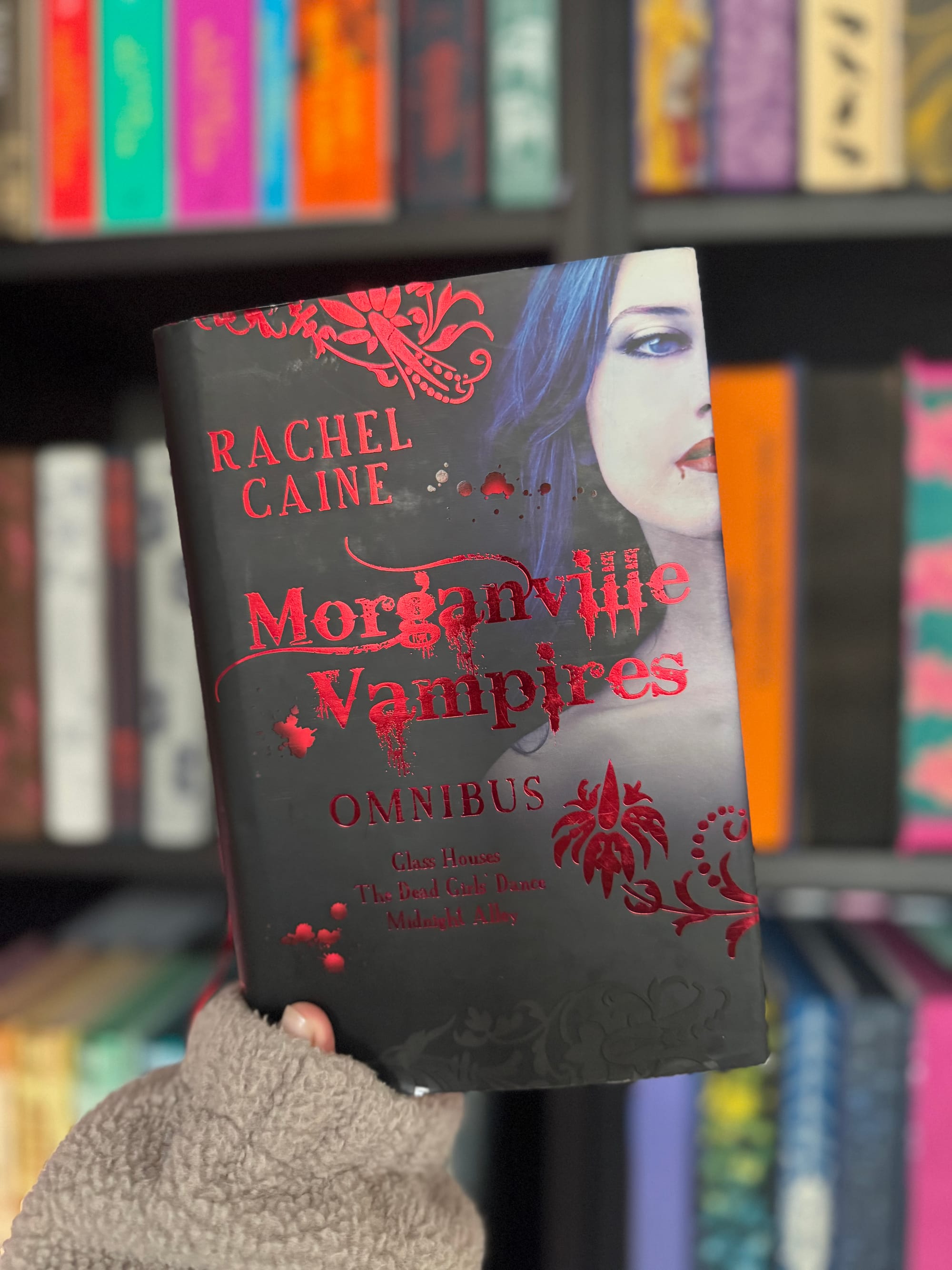 Hand holds "Morganville Vampires Omnibus" by Rachel Caine. Black cover with red text, woman's face. Shelves with colorful books in background.